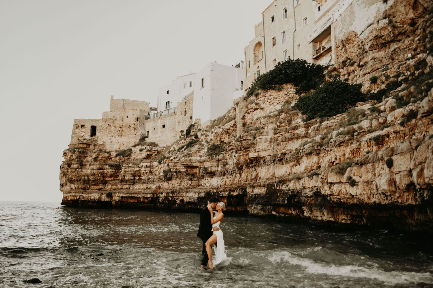 A couple stands in shallow ocean water beneath white cliffside buildings in Italy .