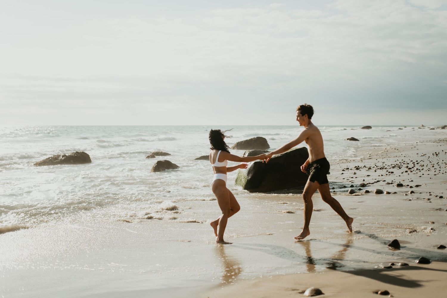 Two people joyfully dancing together on a rocky beach at sunset with waves and stones in the background.