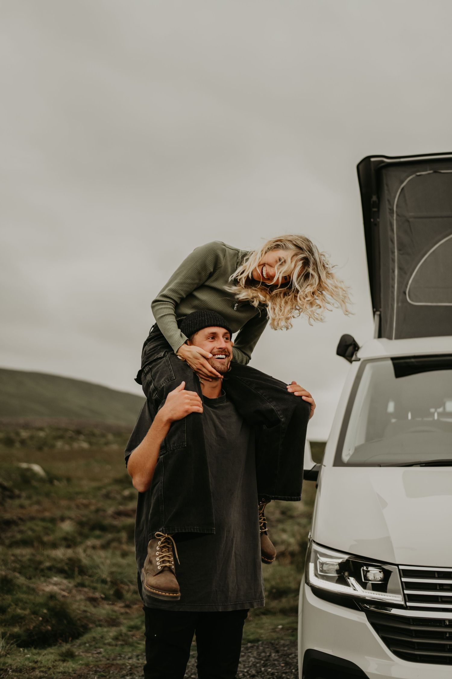 A couple shares a playful moment near their camper van in a remote mountainous landscape on an overcast day.