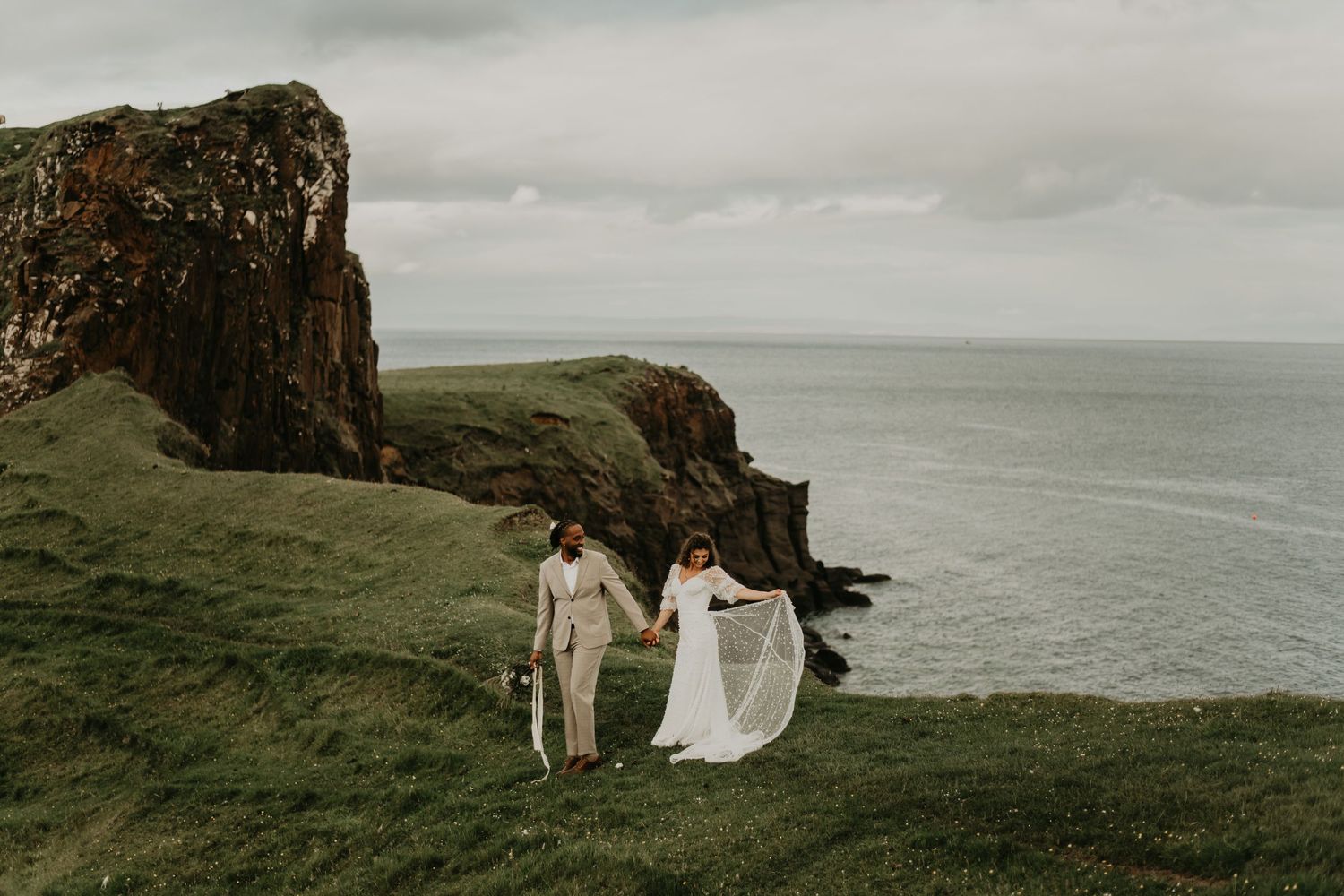 A couple walks along dramatic coastal cliffs overlooking the ocean on their wedding day in isle of Skye Scotland.