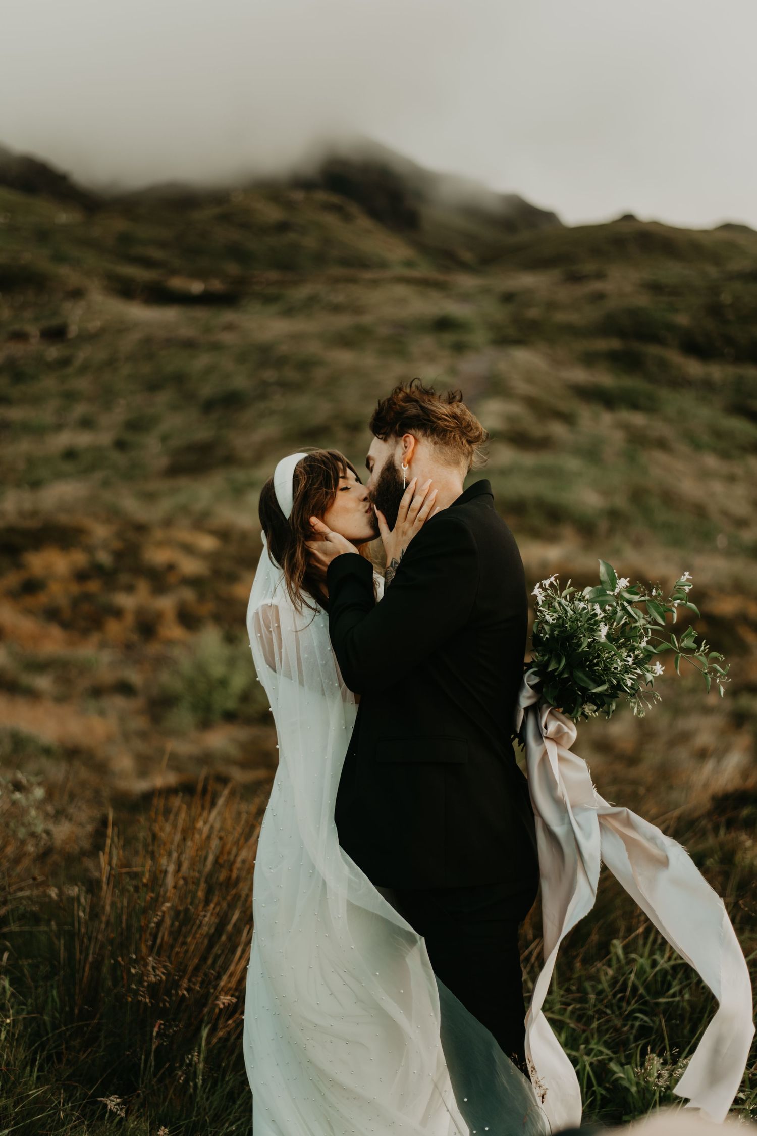 Couple in wedding attire embrace on windswept hillside with bride in flowing white dress holding greenery bouquet.