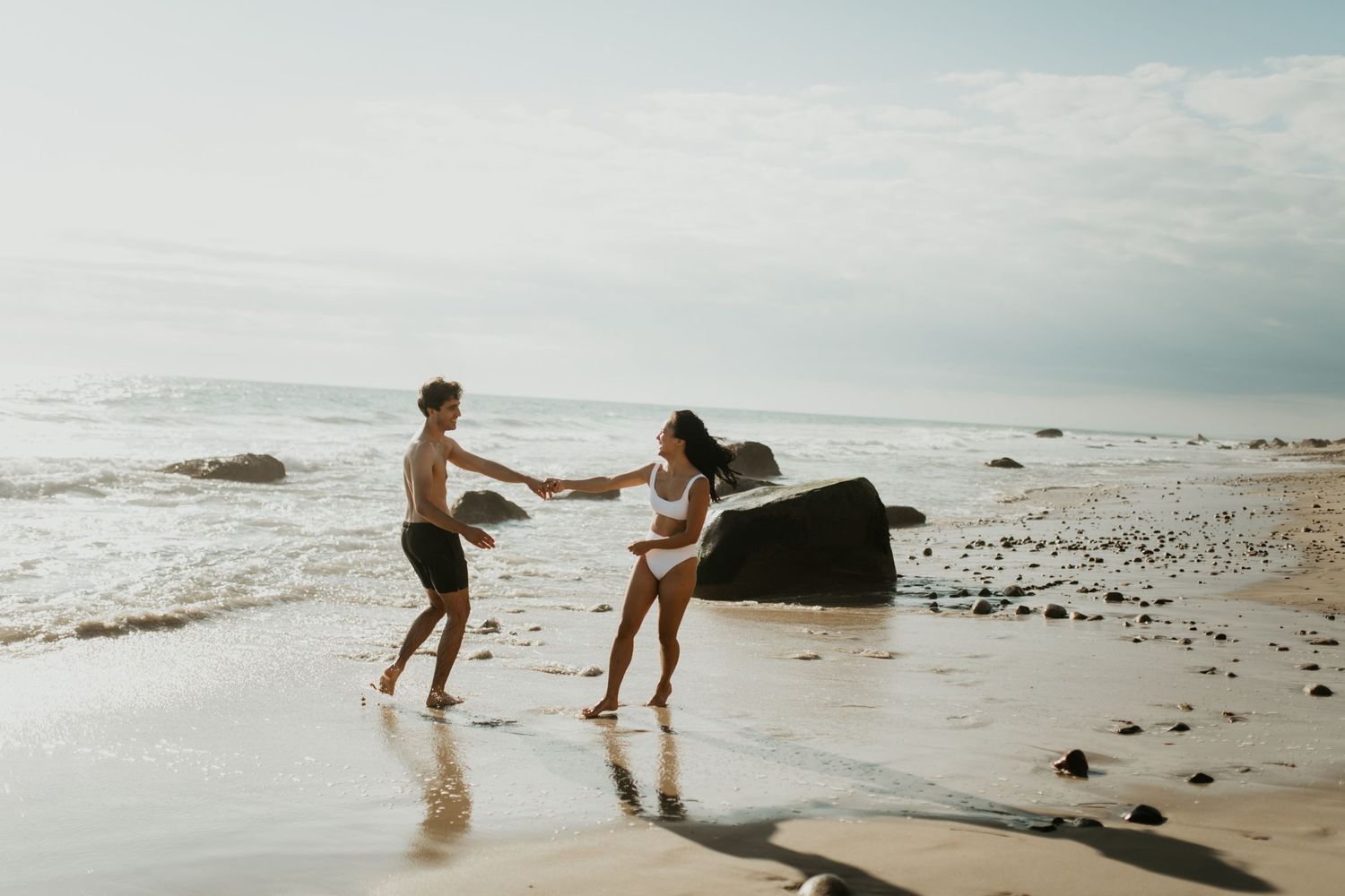 Two people dancing and splashing in the shallow water at a rocky beach during sunset.