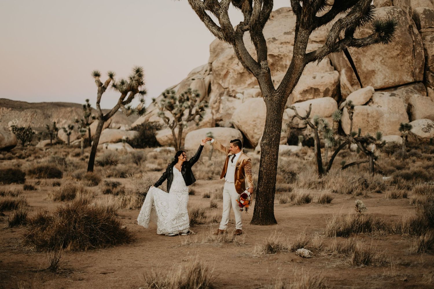 A couple in wedding attire poses among Joshua trees in a desert landscape at sunset.