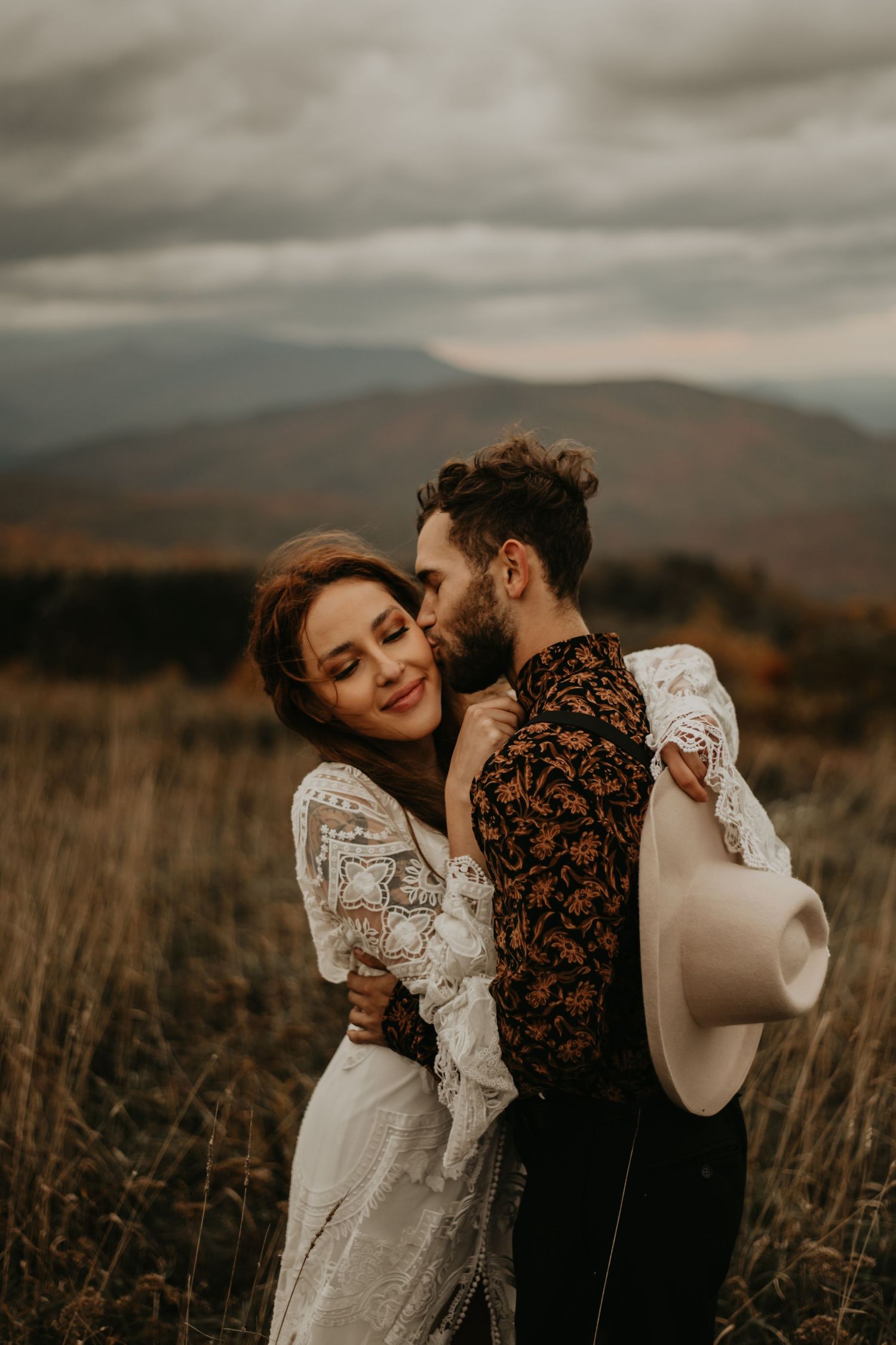 Intimate couple embraces in a field with mountains and dramatic clouds in the background.