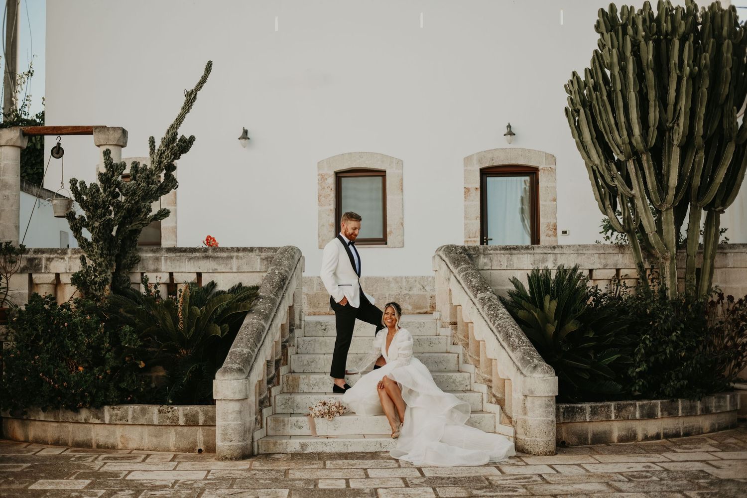 A playful wedding photo on white stone stairs with cacti and Mediterranean architecture.