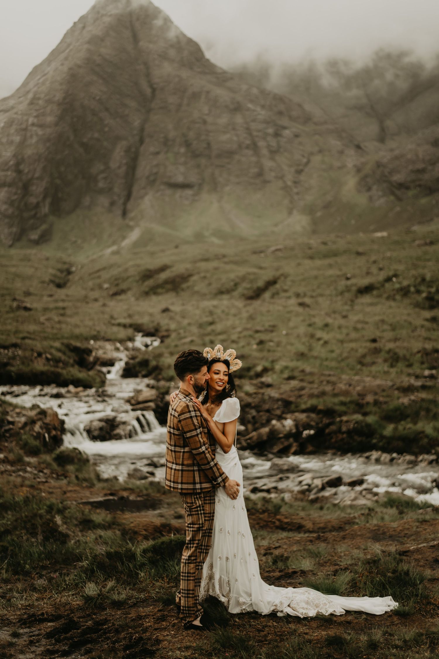 A couple embraces near a mountain stream with dramatic cloudy peaks in the background in isle of Skye Scotland.