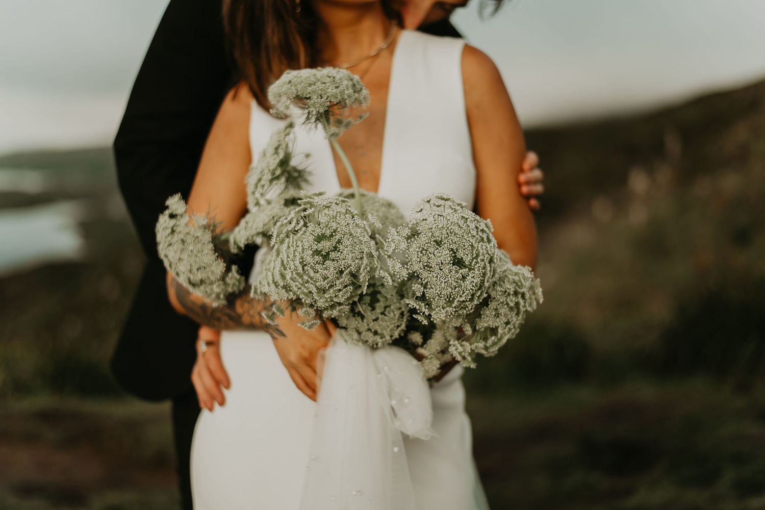 A wedding couple holds white and pale green floral bouquets while posing against a mountainous backdrop.