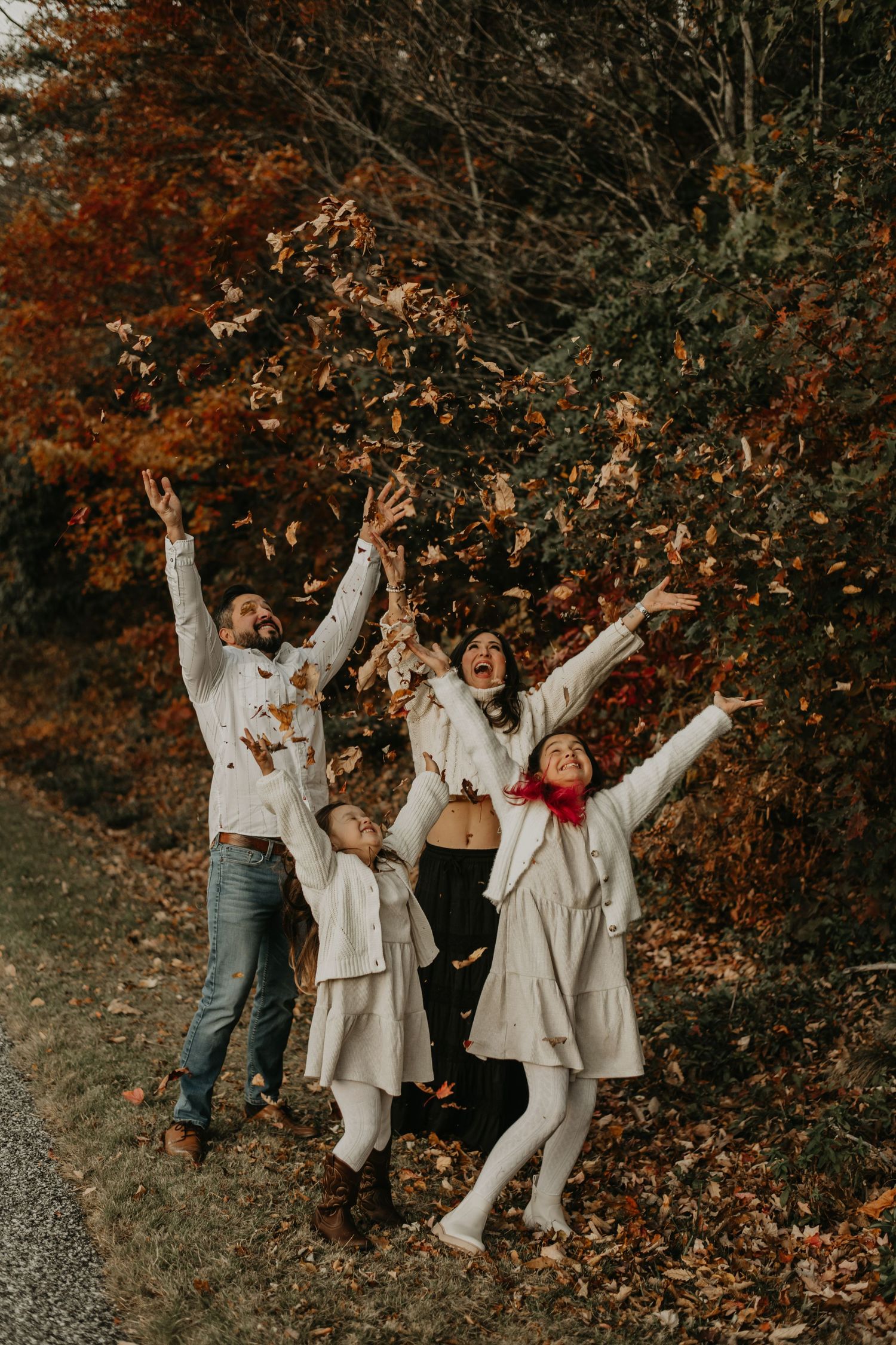 A group celebrates joyfully among autumn foliage wearing white sweaters and casual attire in an outdoor setting.