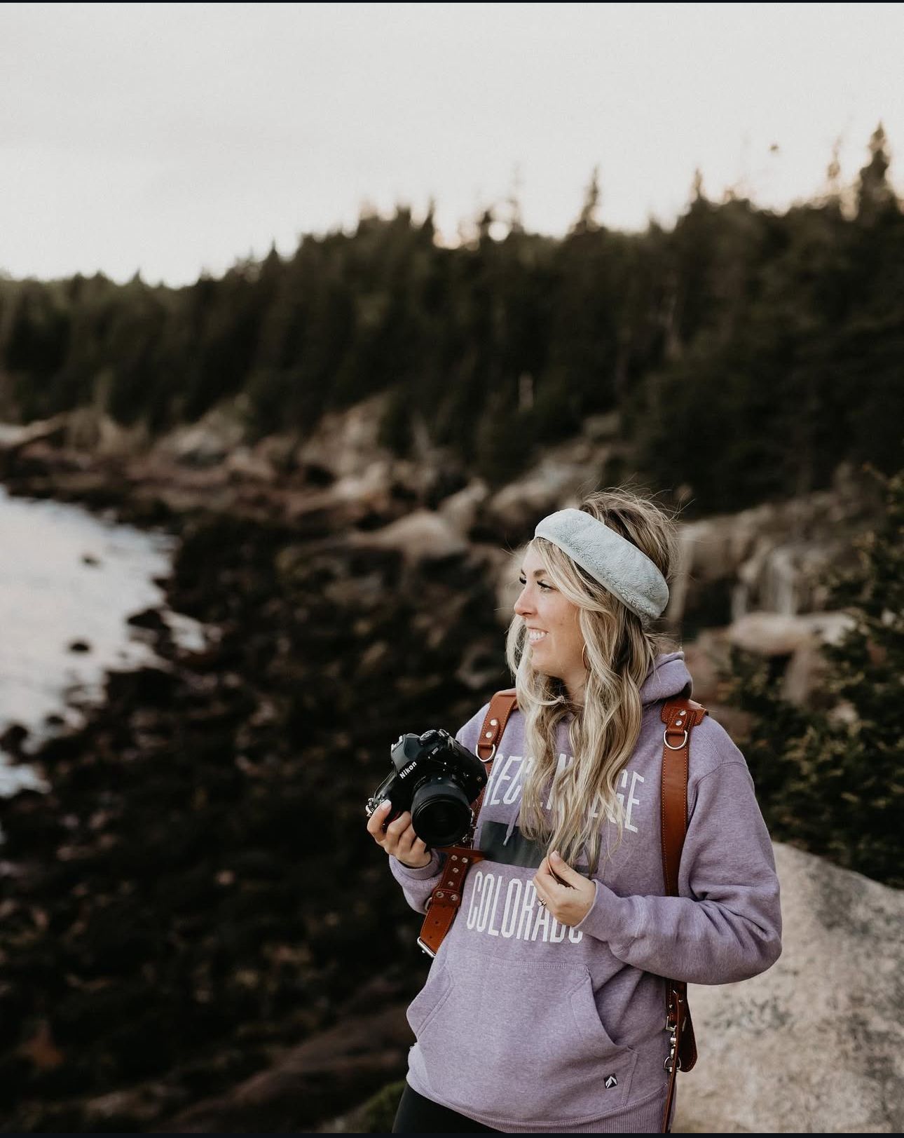 A photographer with a camera enjoys the rugged coastal scenery while wearing a purple sweatshirt and headband.