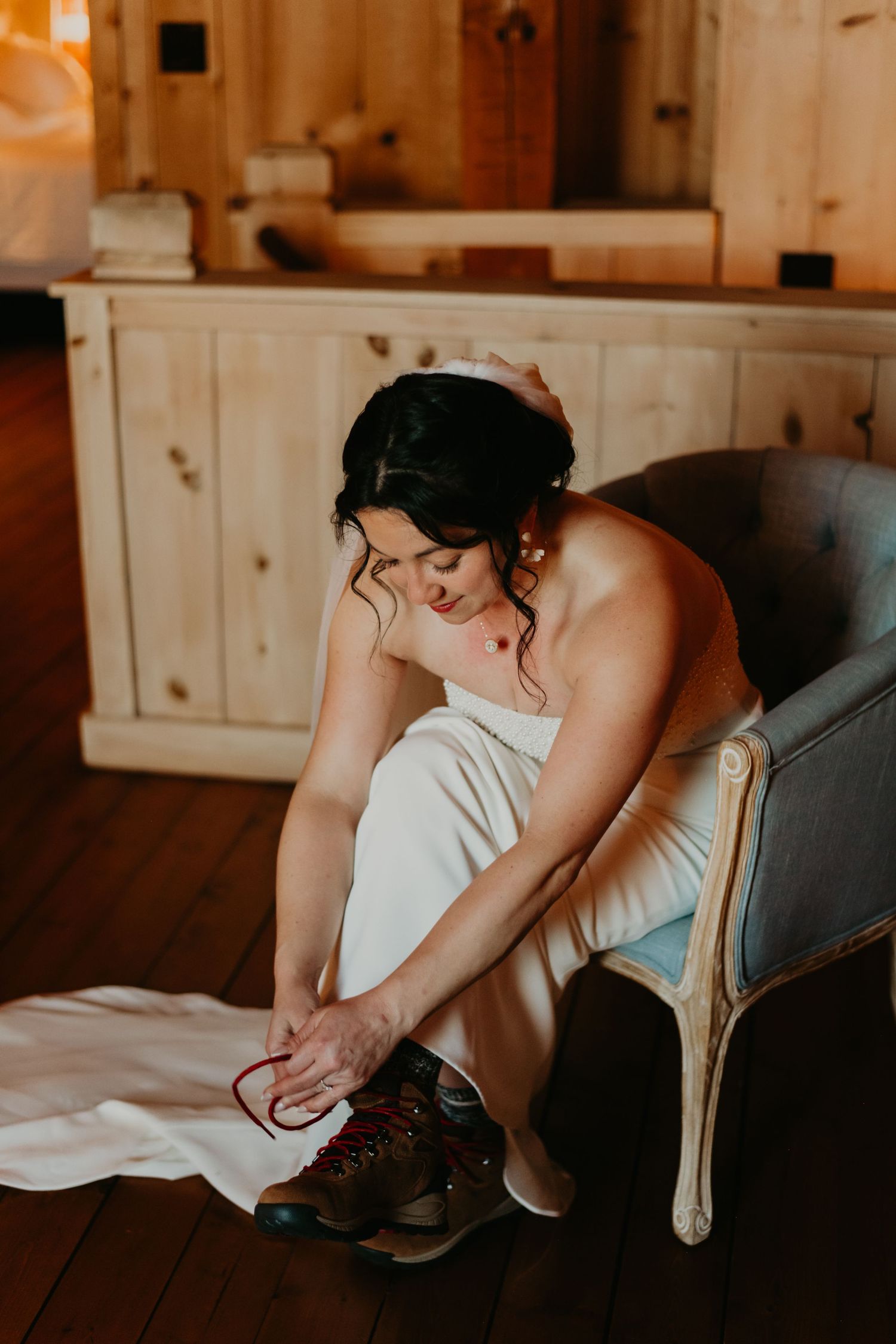 A person in a white dress sits on a vintage chair putting on brown leather boots in warm lighting.