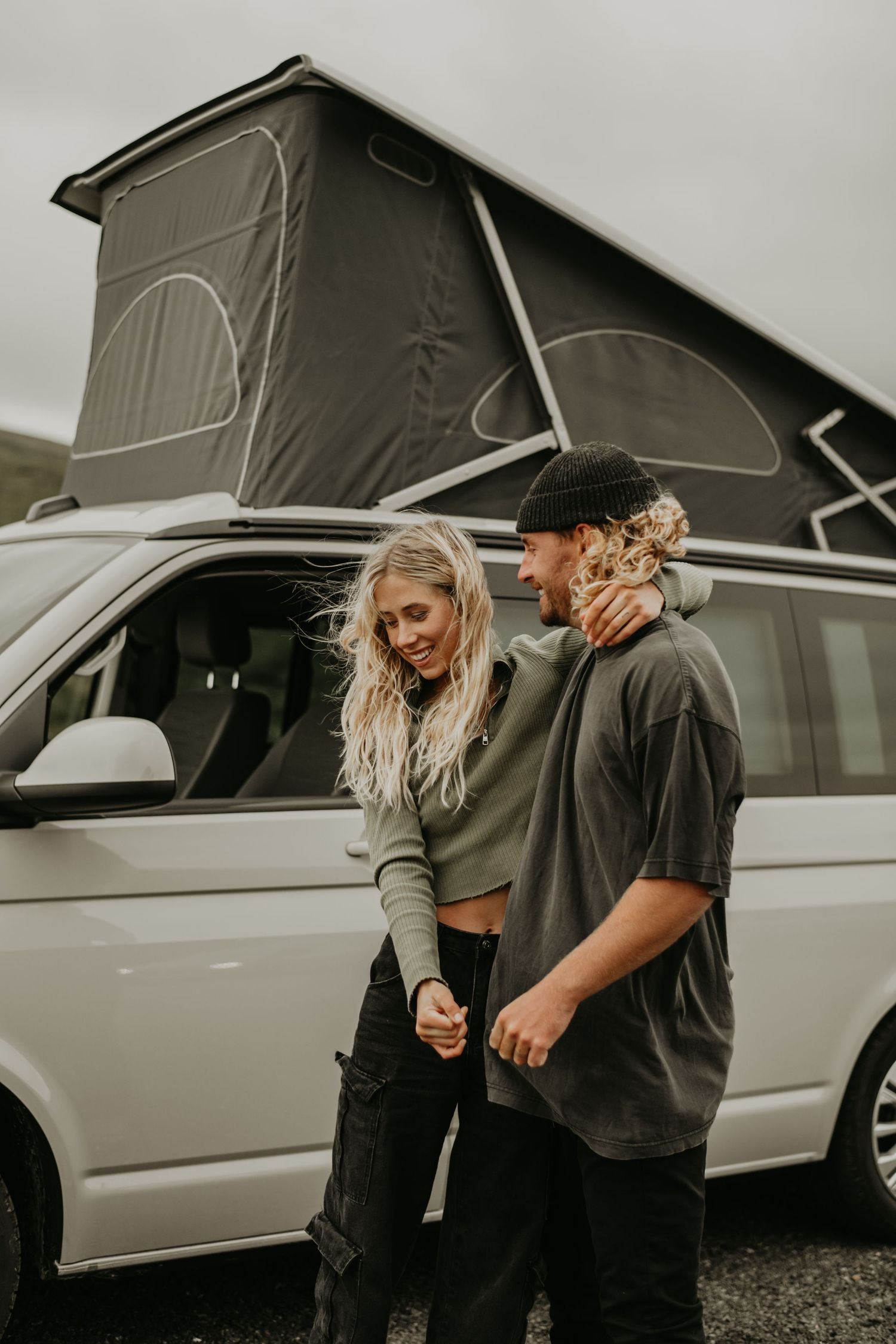A couple sharing intimate candid moments next to a white van with a pop-up roof on a cloudy day.