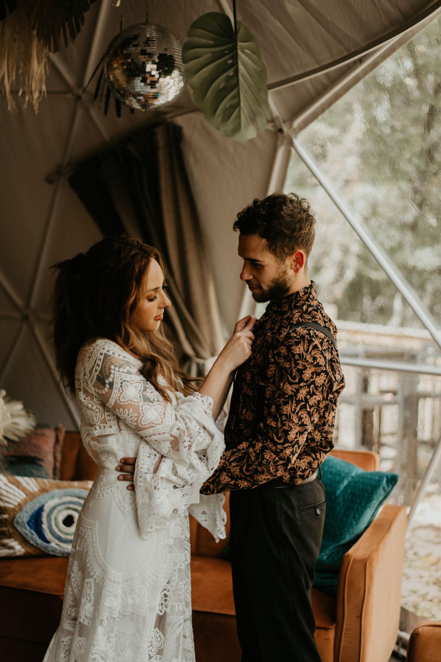 Couple sharing an intimate moment in a boho-styled tent with the bride wearing a lace dress and groom in floral shirt.