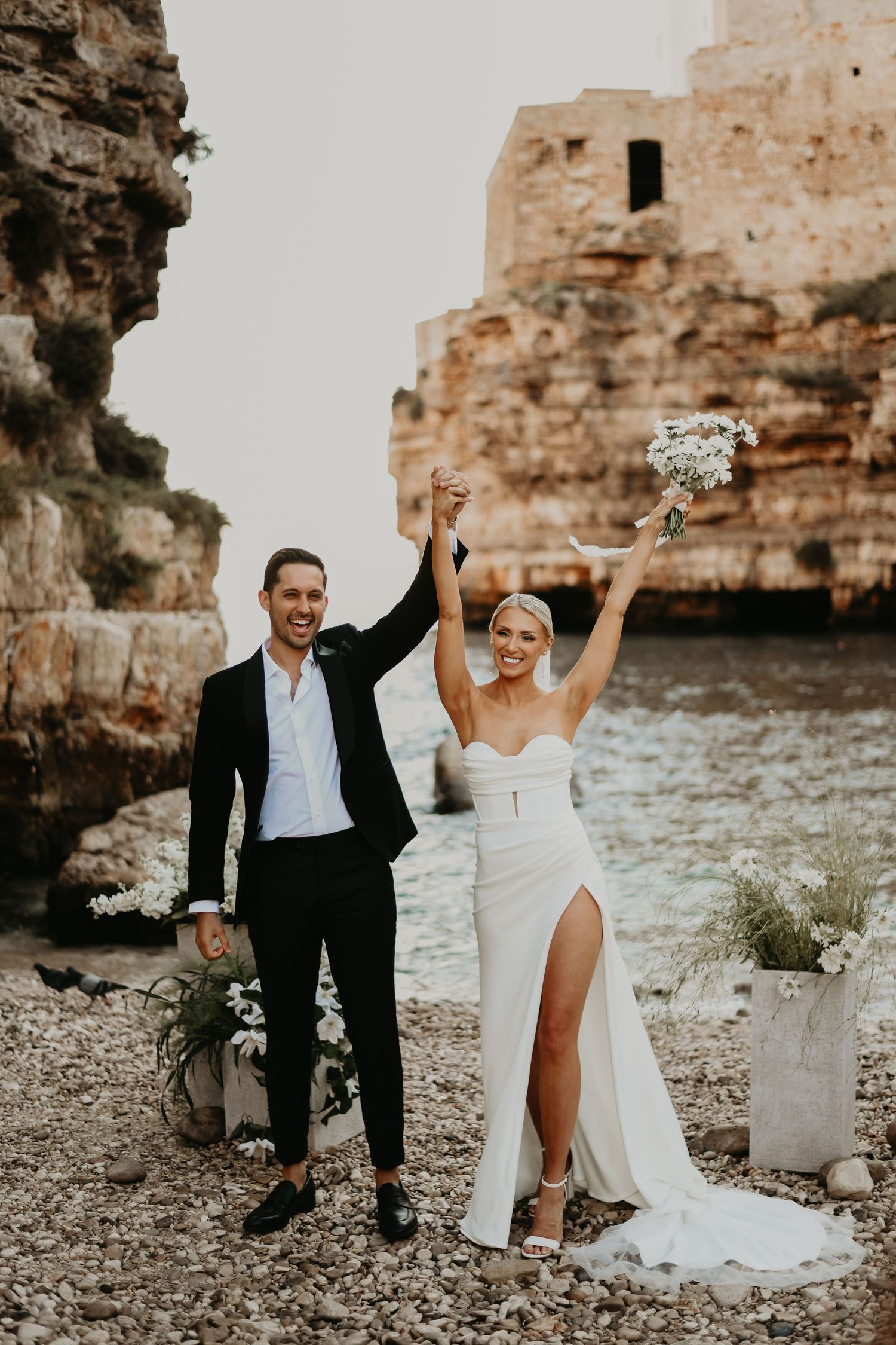 Newlyweds celebrate with raised arms on rocky Mediterranean beach with historic fortress backdrop in Italy.