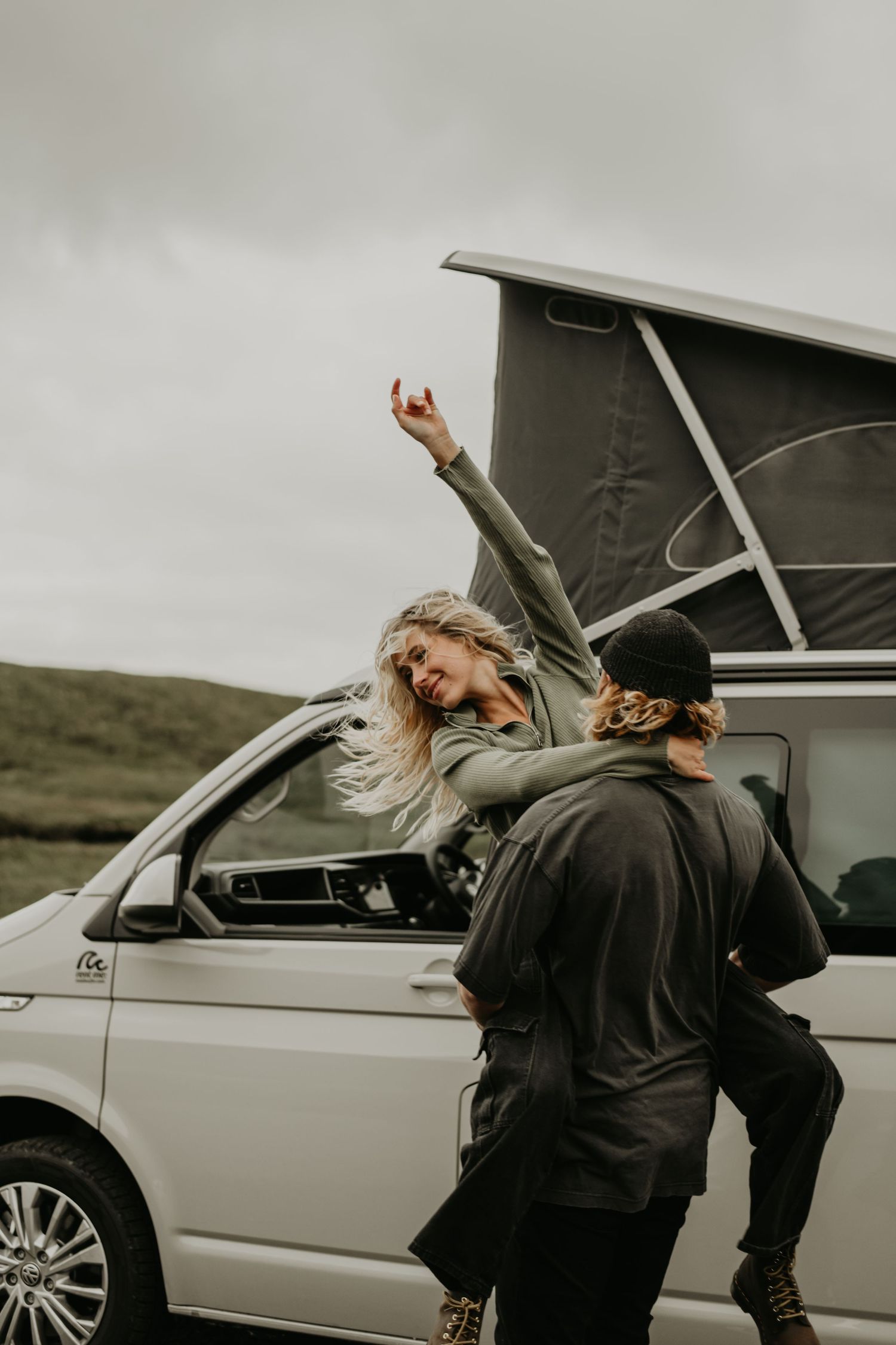 A couple enjoys freedom near their camper van on a cloudy day with one person raising their hand through the roof.