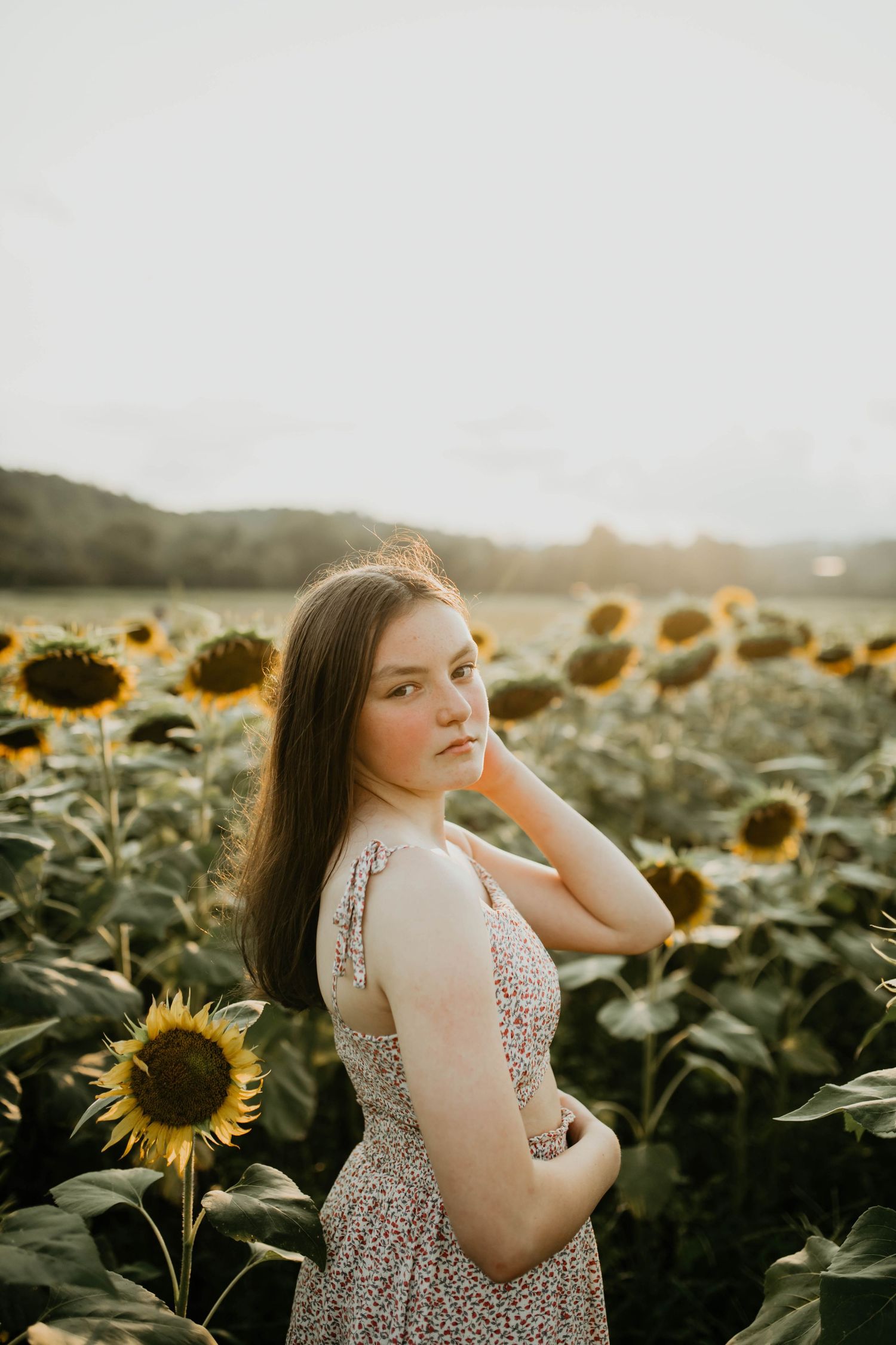 A person poses gracefully in a sunflower field at sunset wearing a floral dress while golden light illuminates the scene.
