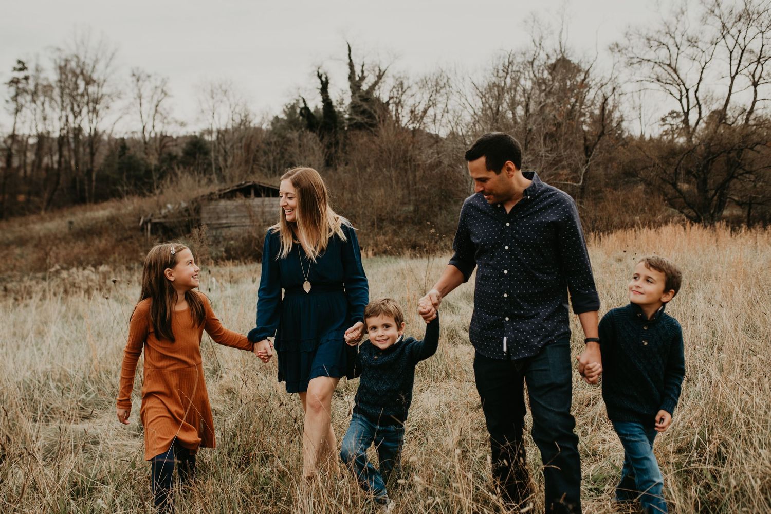 A family group holding hands walks through a golden tall grass field at sunset in autumn.