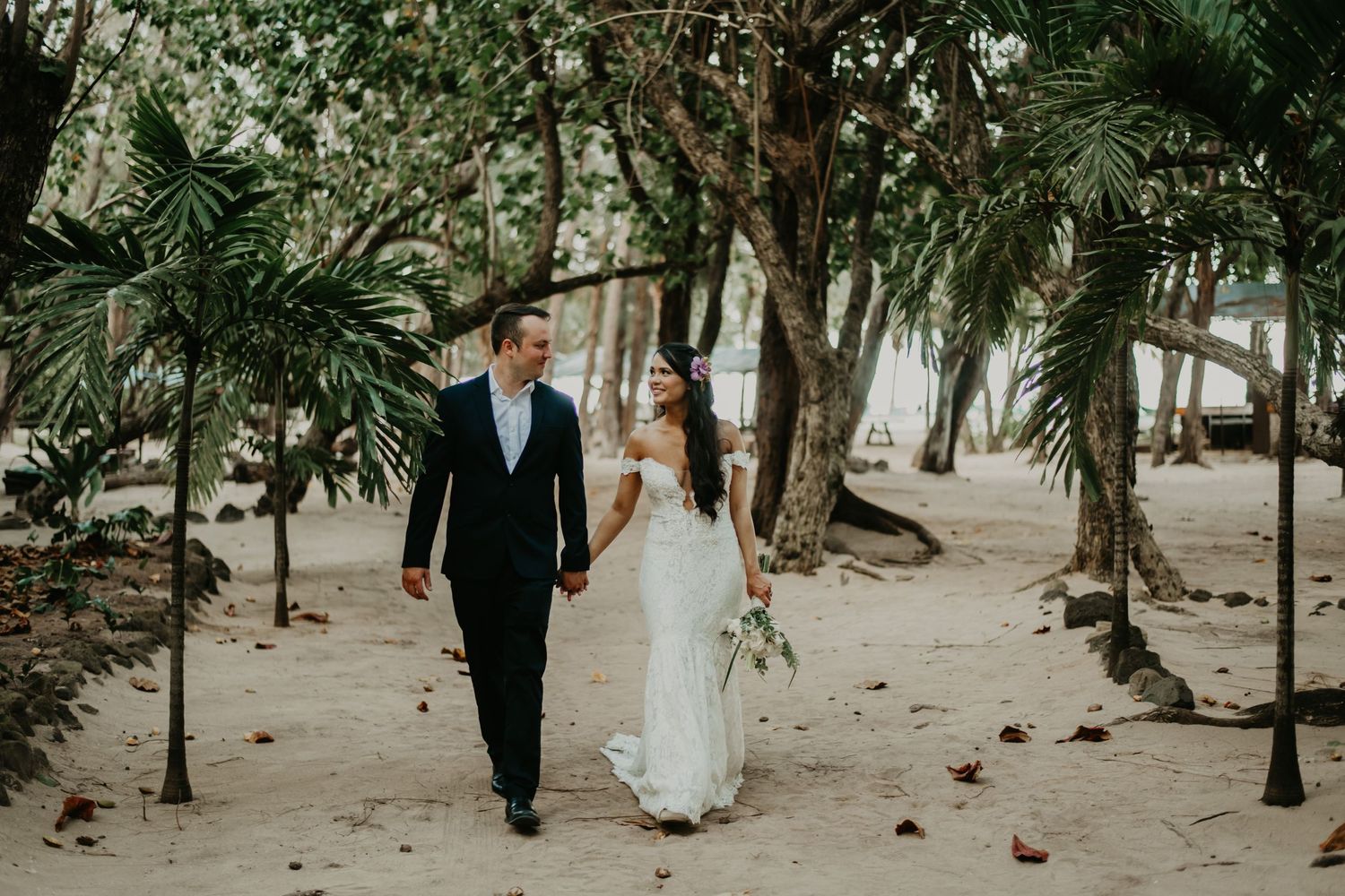 Newlyweds walk hand in hand through a tropical palm tree lined beach path in Hawaii.
