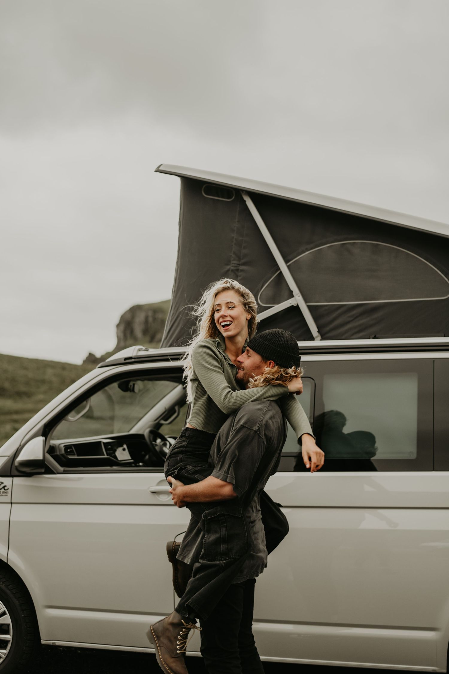 A joyful couple embraces beside a silver camper van with pop-up roof on a cloudy day in the countryside.