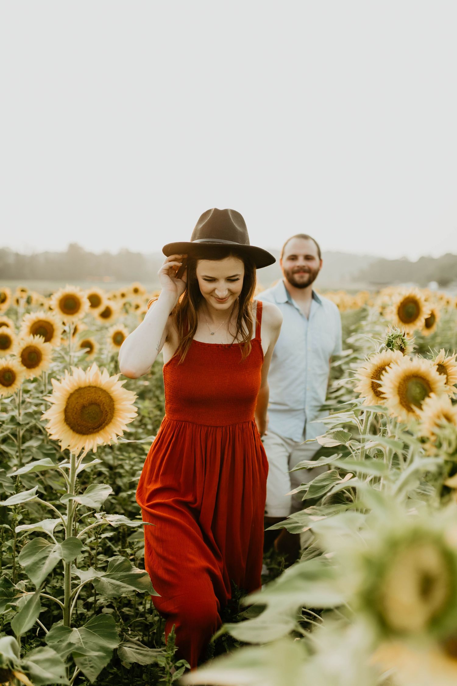 Couple walking through blooming sunflower field with sunlight, woman wearing red dress and brown hat.