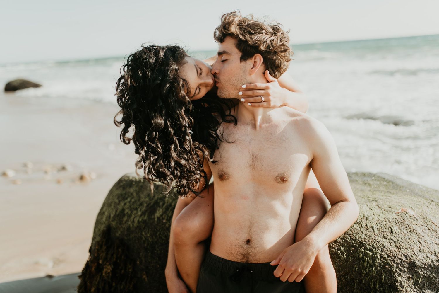 Two people share an intimate moment on a rocky beach at sunset with ocean waves in the background.