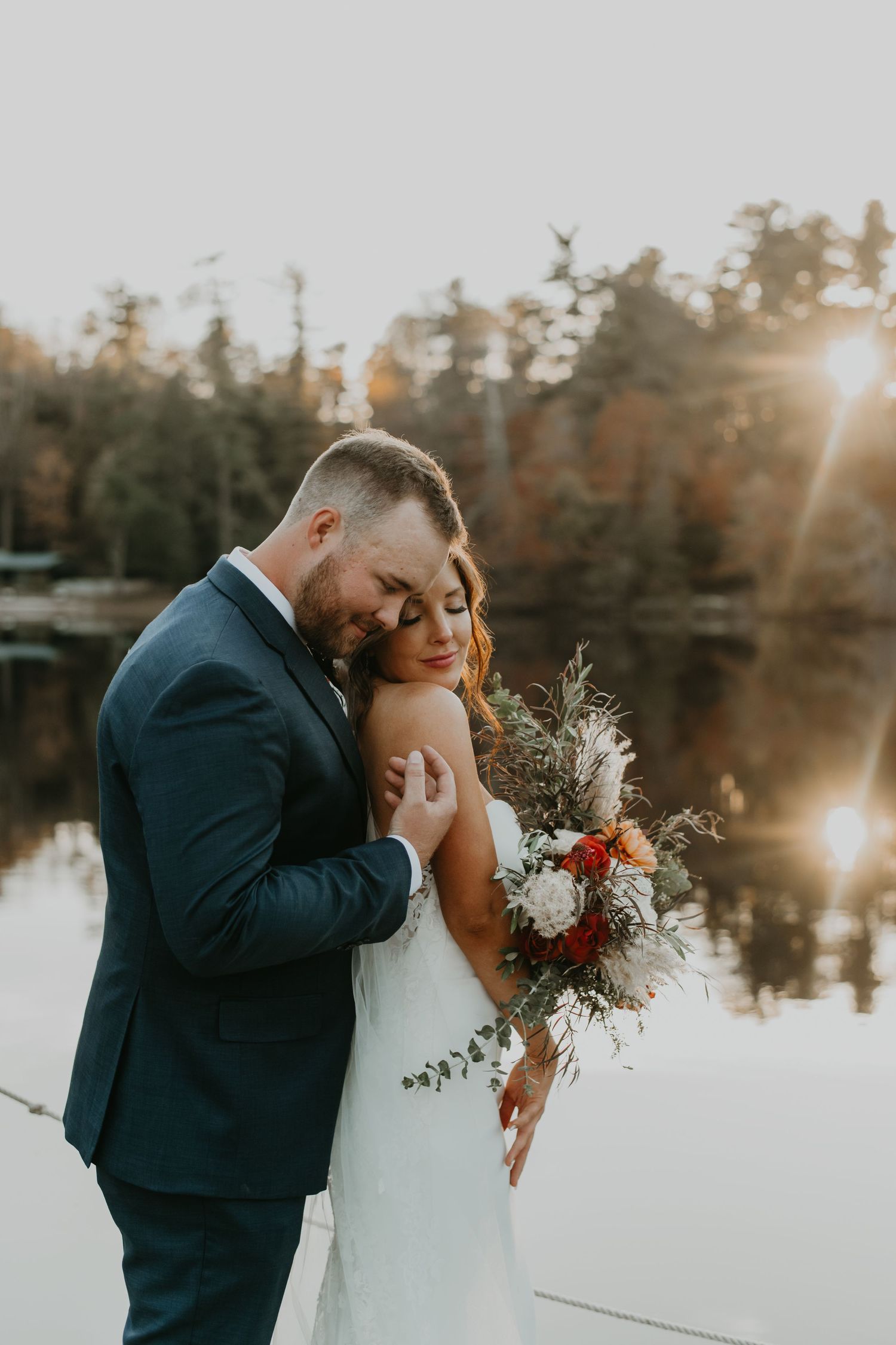 Romantic wedding couple embraces by a lake at sunset with the bride holding a rustic bouquet in North Carolina.