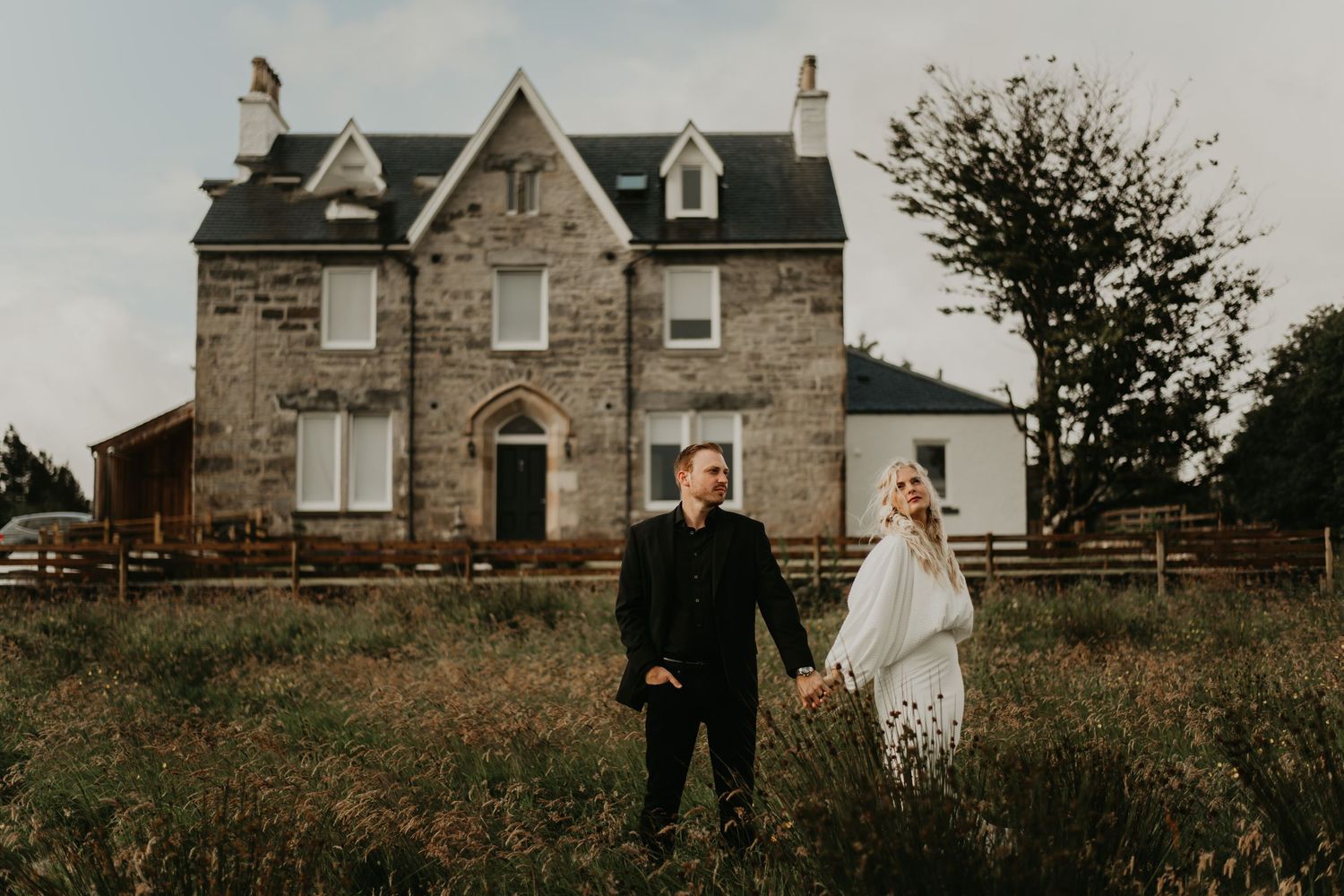 A wedding couple stands in front of a stone house with dormers and climbing vines on a moody autumn day in isle of skye Scotland.