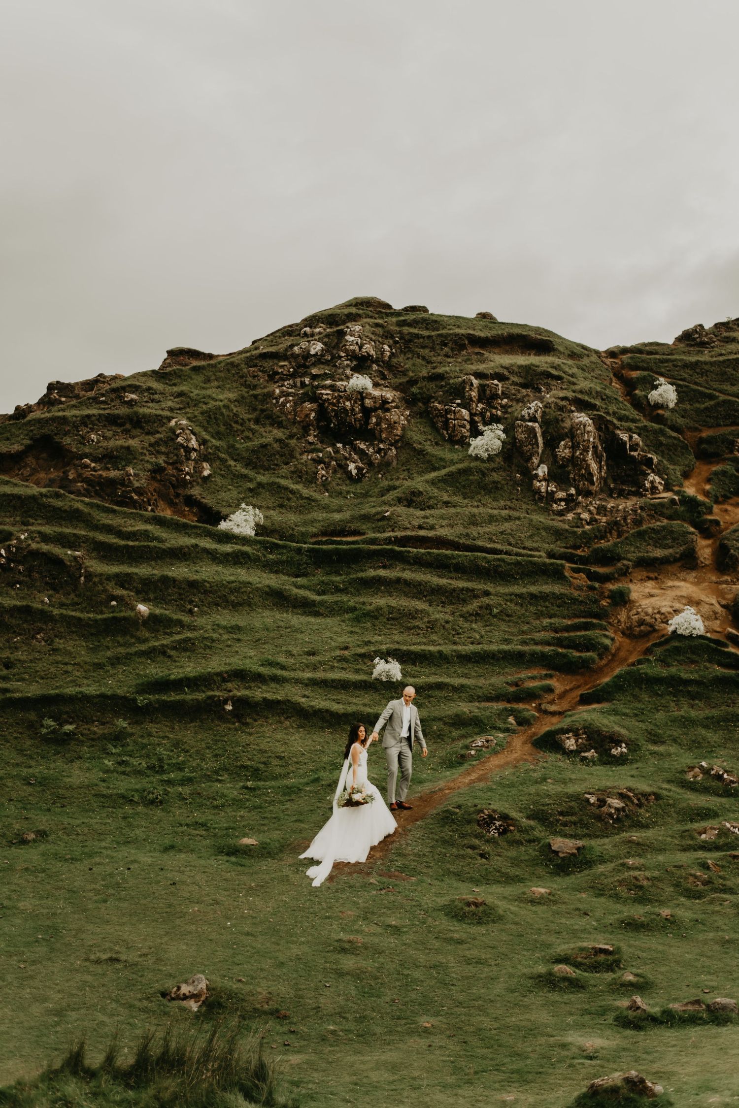 Couple in wedding attire walking up winding path on dramatic green hillside.