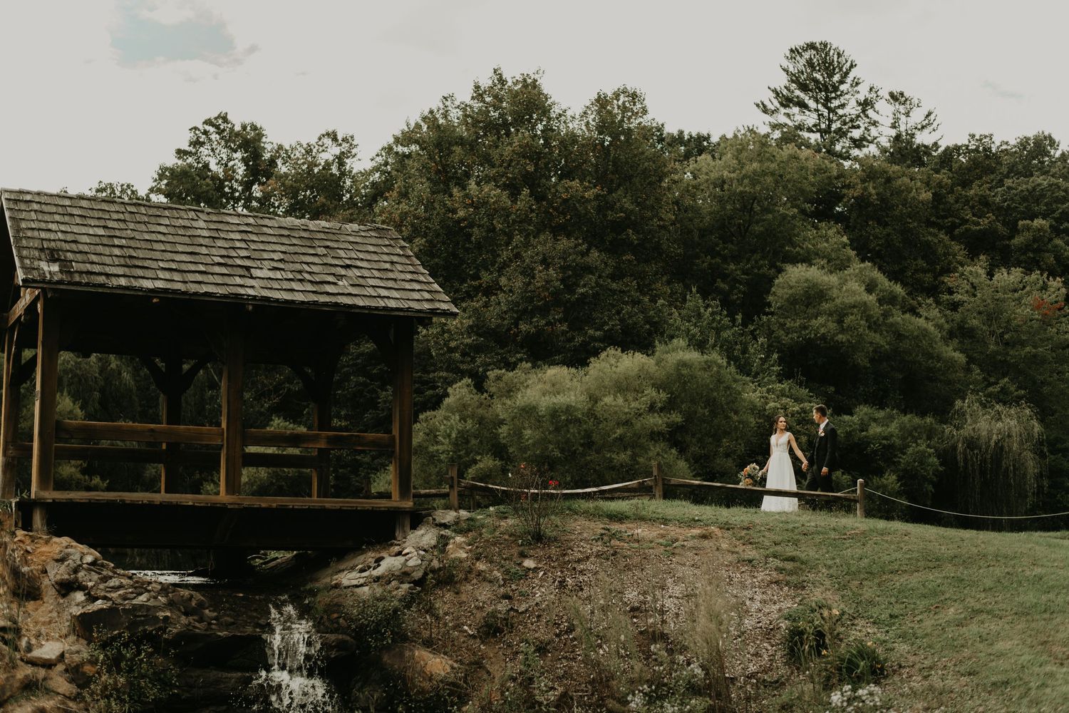 A distant view of a rustic wedding venue with a covered bridge and waterfall in Asheville north Carolina.