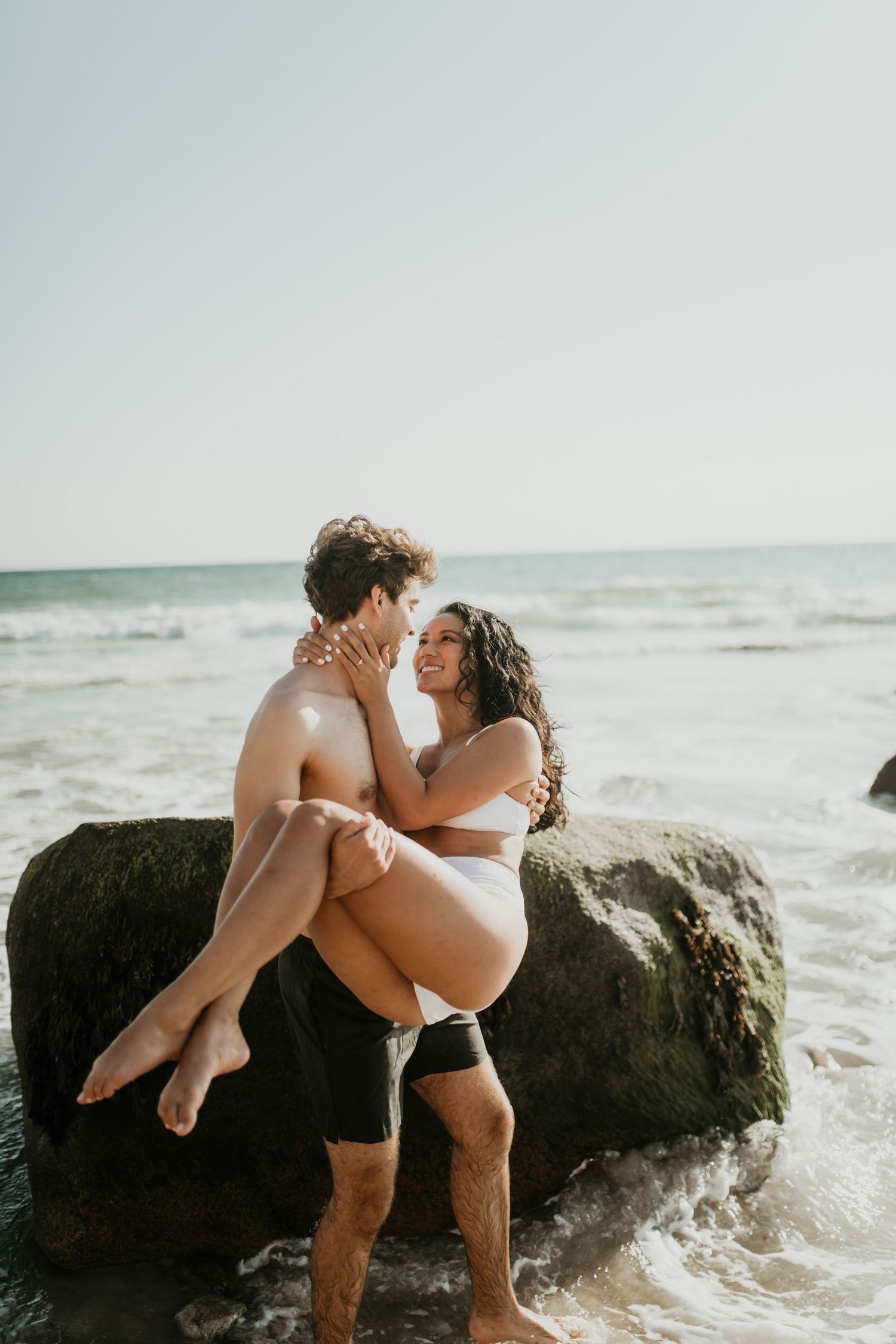 A romantic beach couple embrace on a large rock by the ocean shore at sunset.