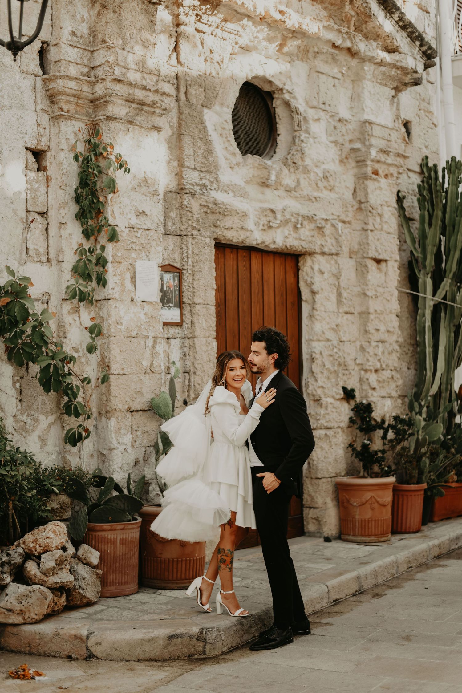 Stylish wedding couple poses by vintage stone building entrance with potted cacti in Italy.