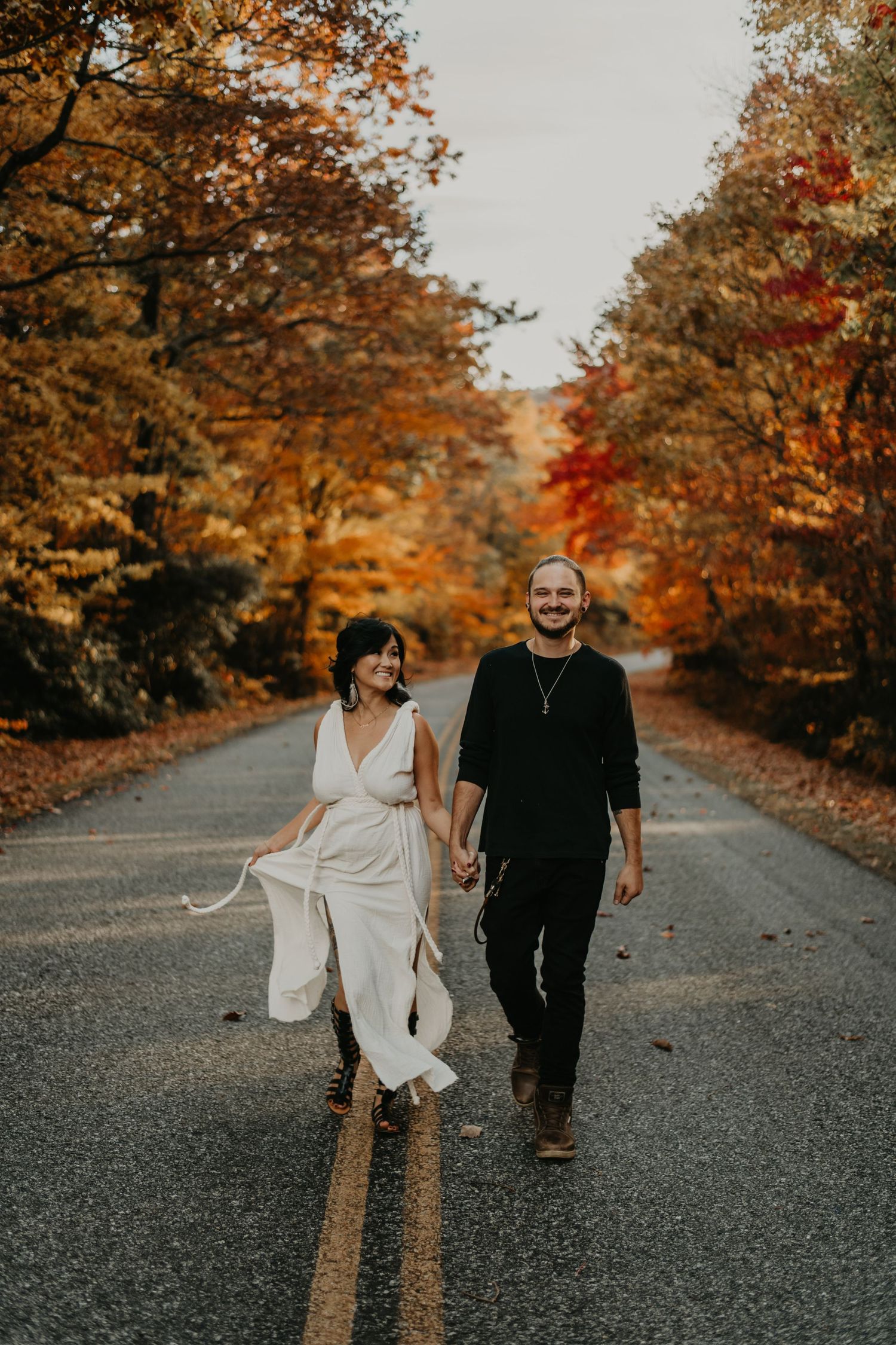 A couple strolls down a gravel road lined with vibrant autumn trees displaying orange and red fall foliage.