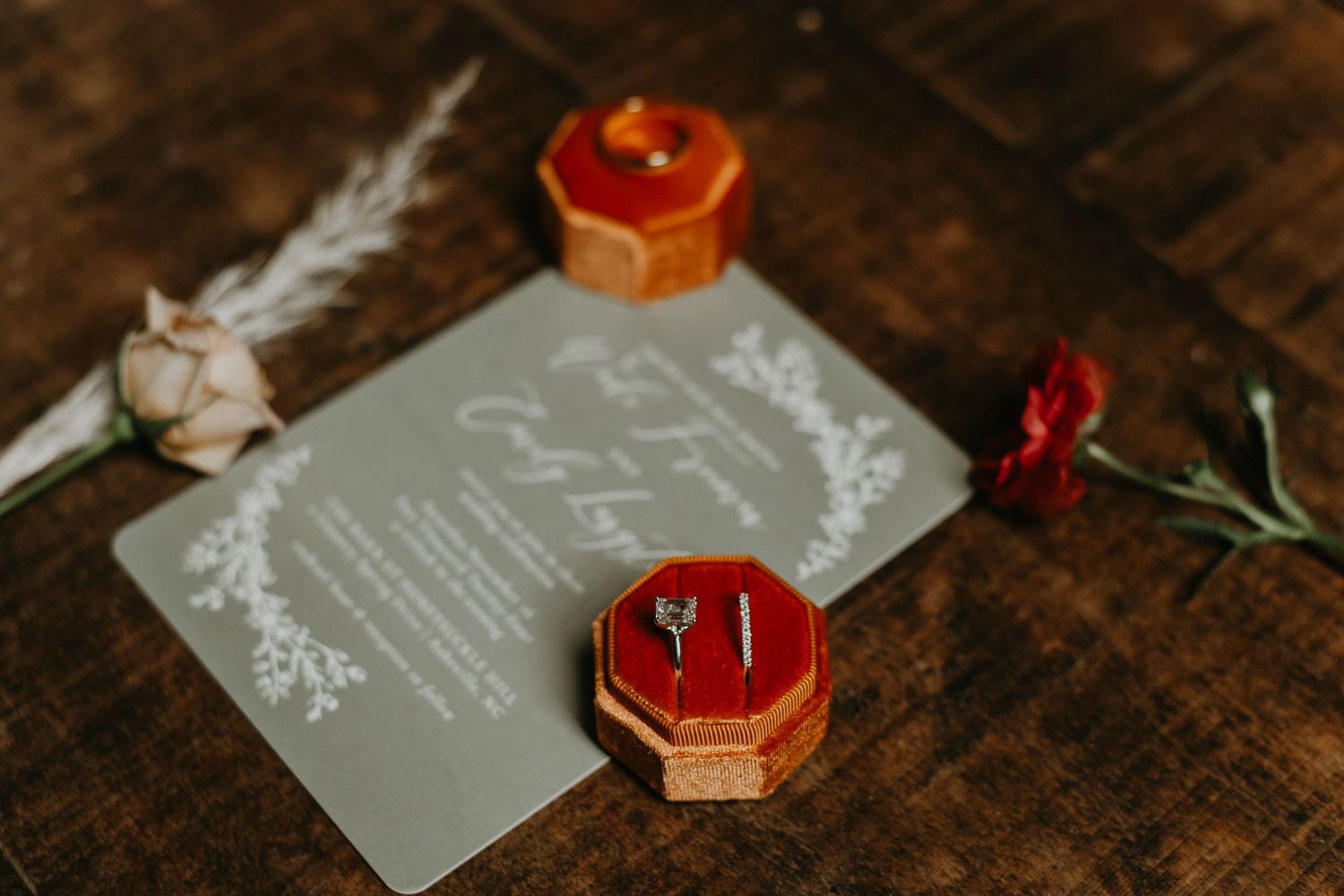Wedding rings in red velvet hexagonal boxes displayed with an invitation on rustic wooden table.