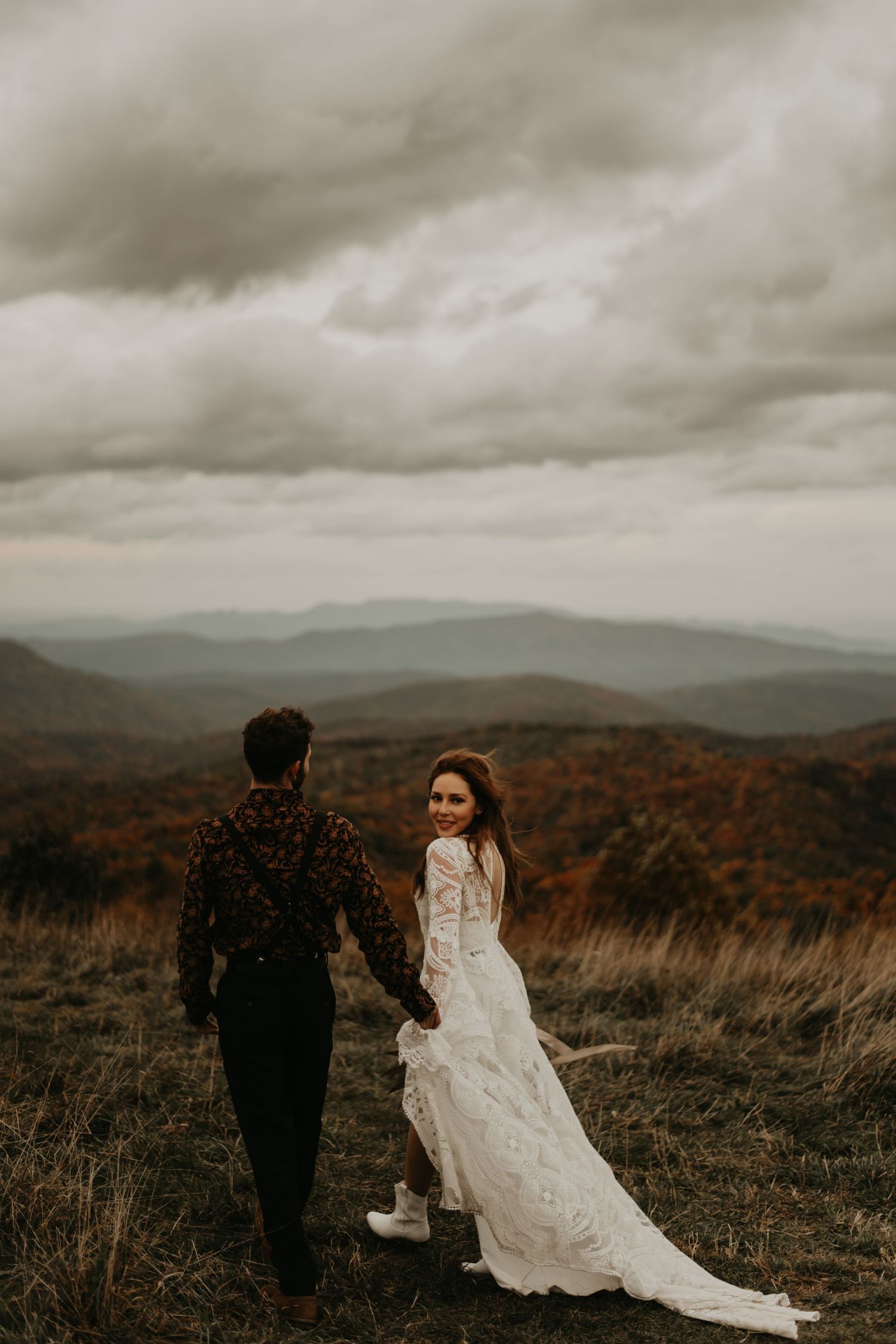 A couple in formal wear stands on a hilltop with a dramatic cloudy mountain landscape behind them in North Carolina.