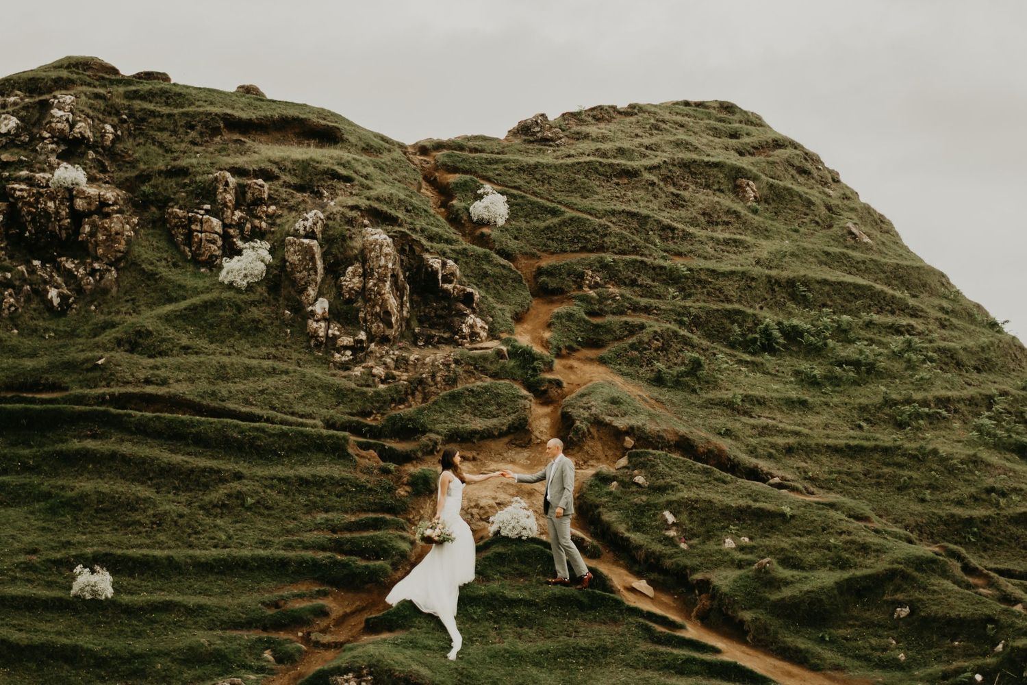 A couple in wedding attire walks along a winding dirt path on a green mountainside in isle of Skye Scotland.