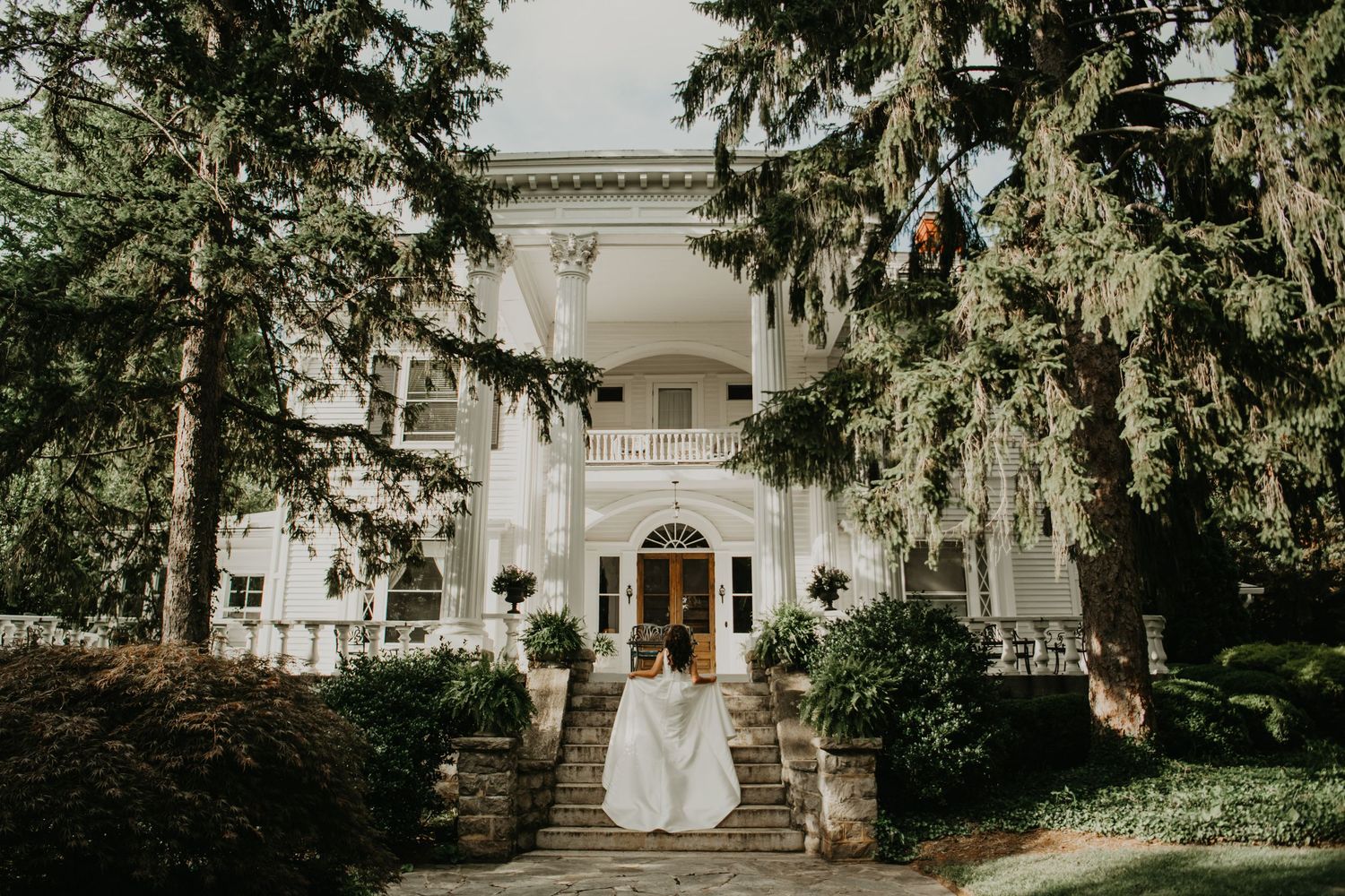 A grand white mansion with stone steps and tall trees serves as an elegant wedding venue backdrop in Asheville, North Carolina.