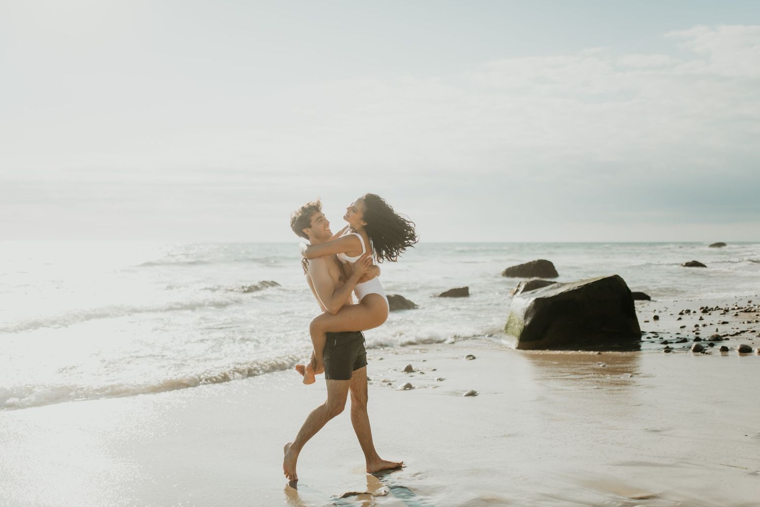 Couple embraces romantically on sandy beach with ocean waves and rocks in the background at sunset.