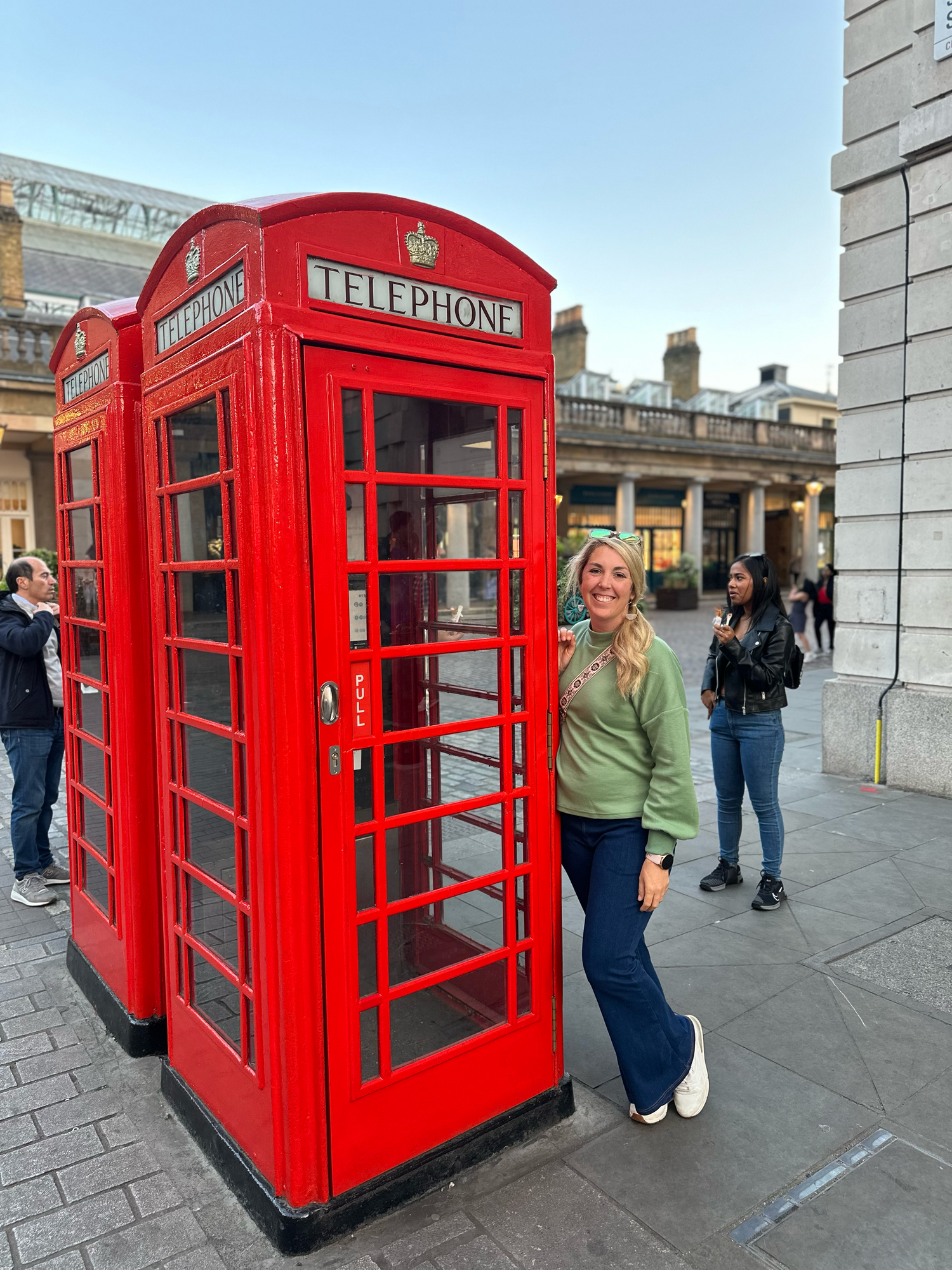 Woman posing next to an iconic red British phone booth on a city street.