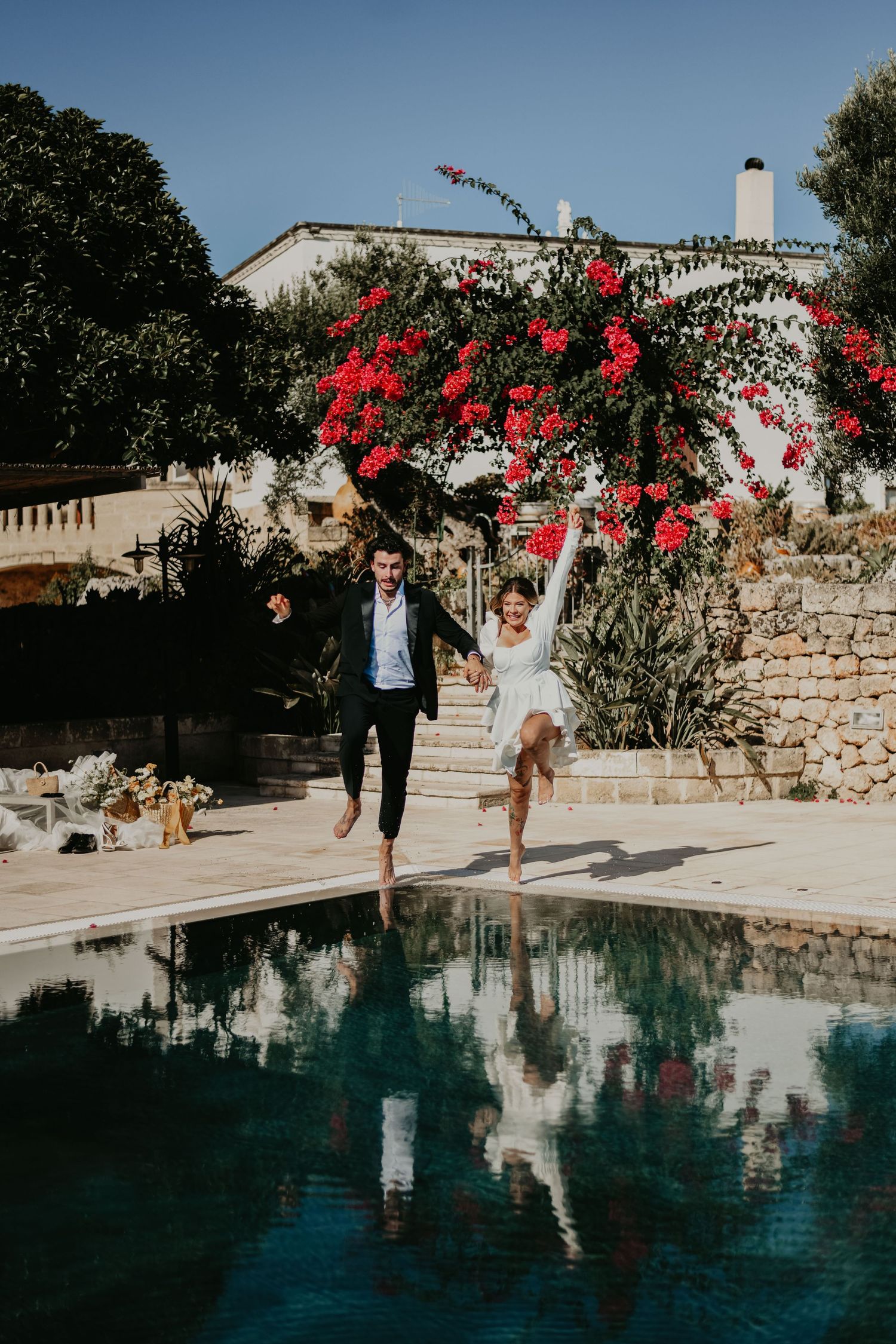 Couple jumping into pool with vibrant red bougainvillea and white villa in background in Puglia Italy.