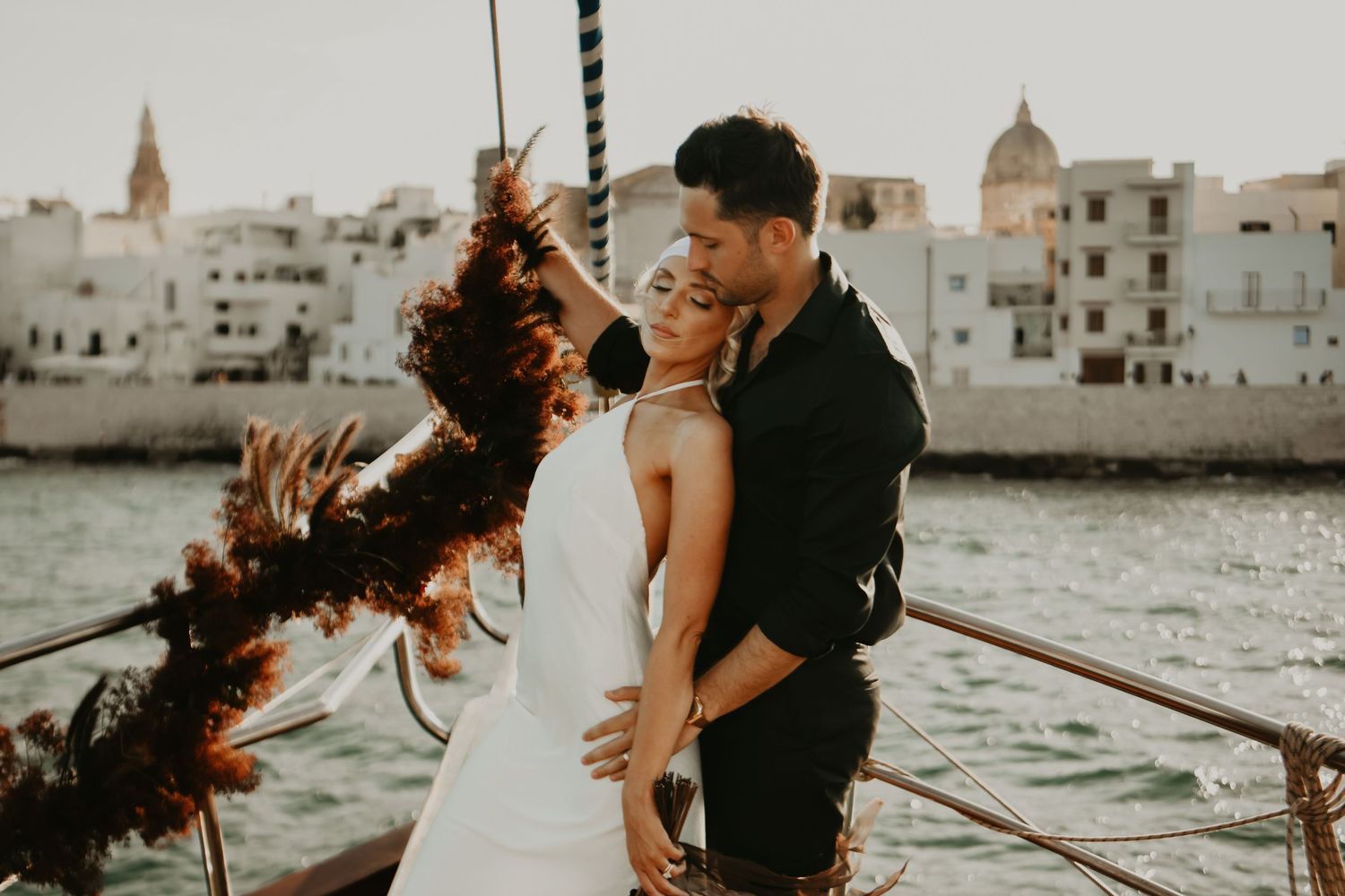 Newlyweds celebrating on boat with colorful smoke bombs against coastal cityscape in Italy.