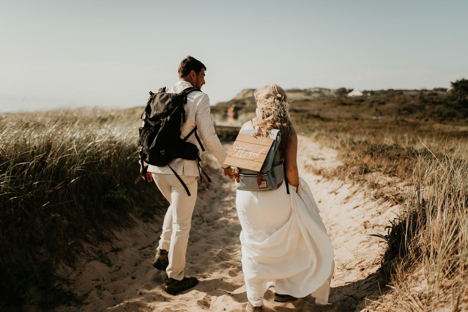 A couple walks hand-in-hand along a sunlit coastal trail through golden grass in Cape Cod.