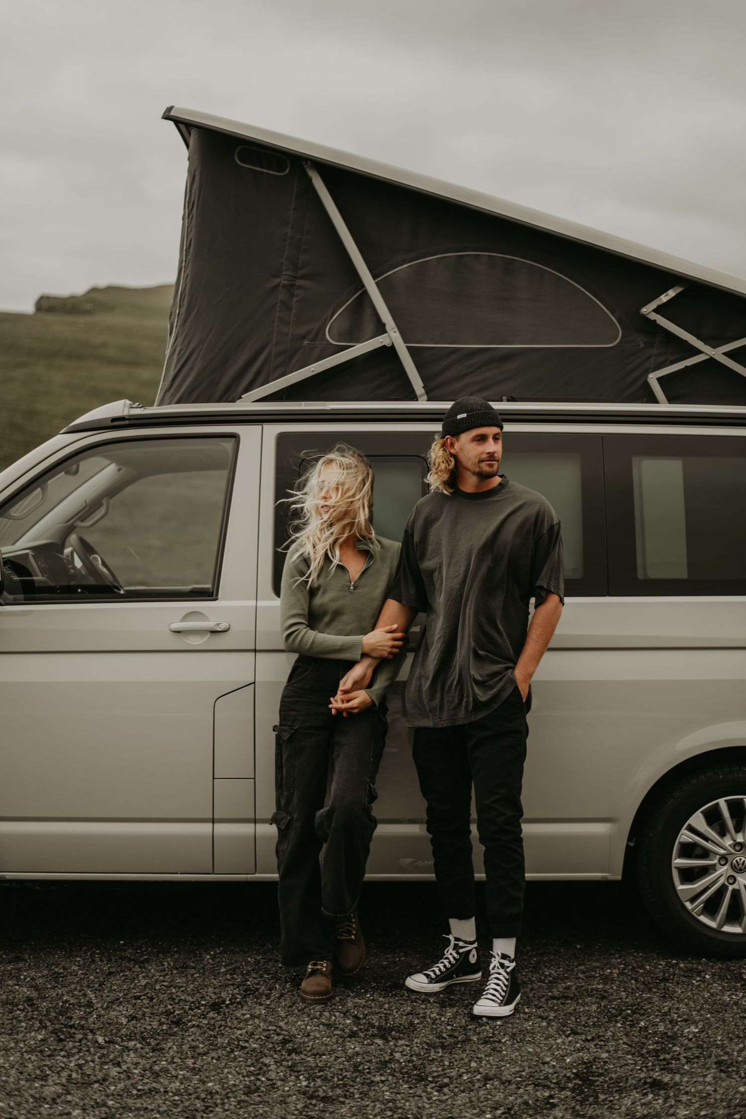 Two people stand beside a silver camper van with pop-up roof on a cloudy day in rugged terrain.