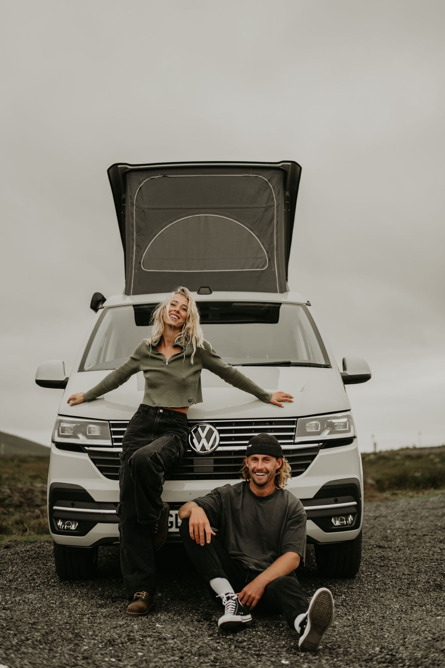 Black and white photo series showing two people posing playfully in front of a white Volkswagen vehicle on a gravel road.