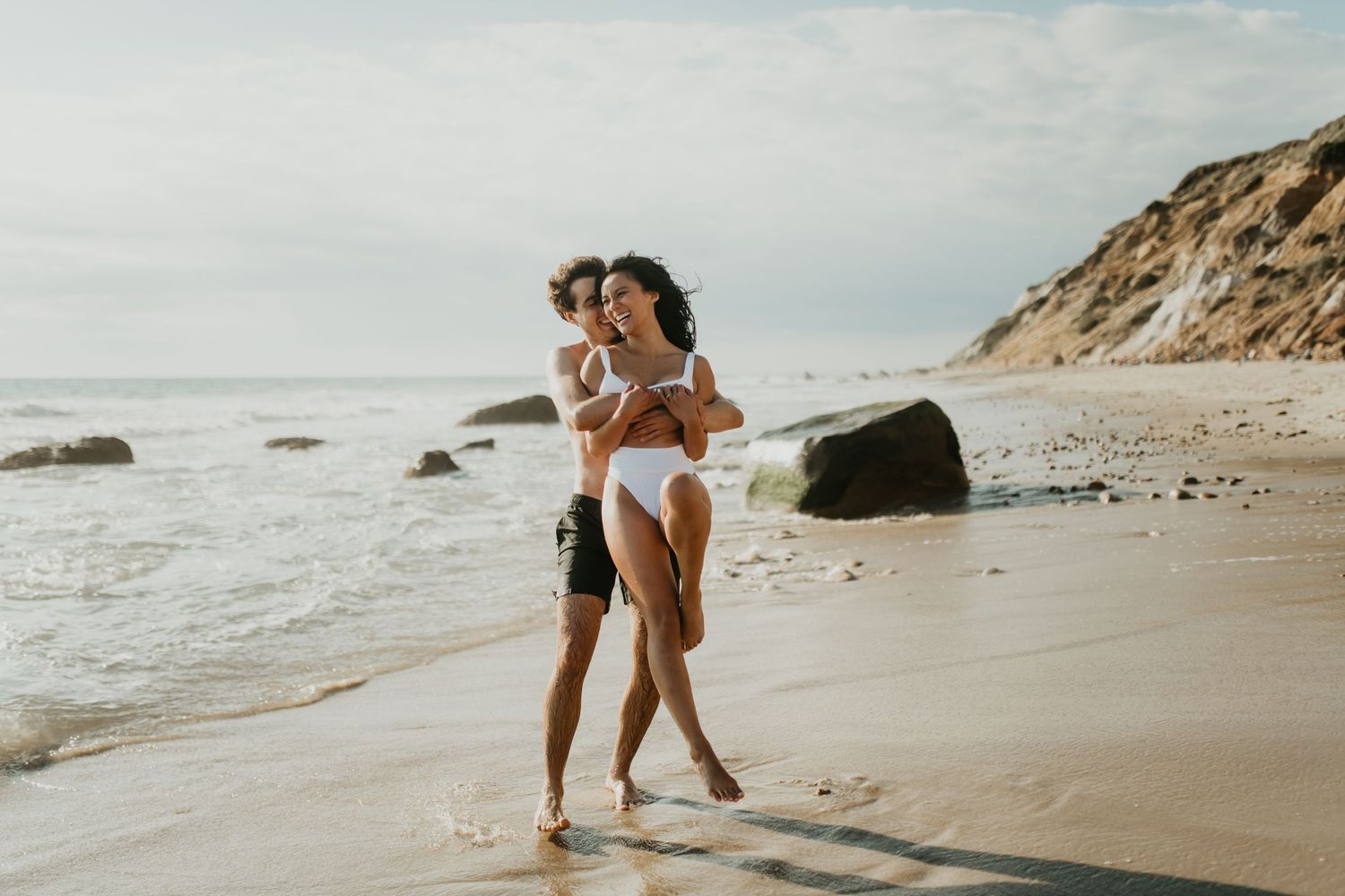 A couple embraces while walking along a sandy beach with cliffs and ocean waves in the background.