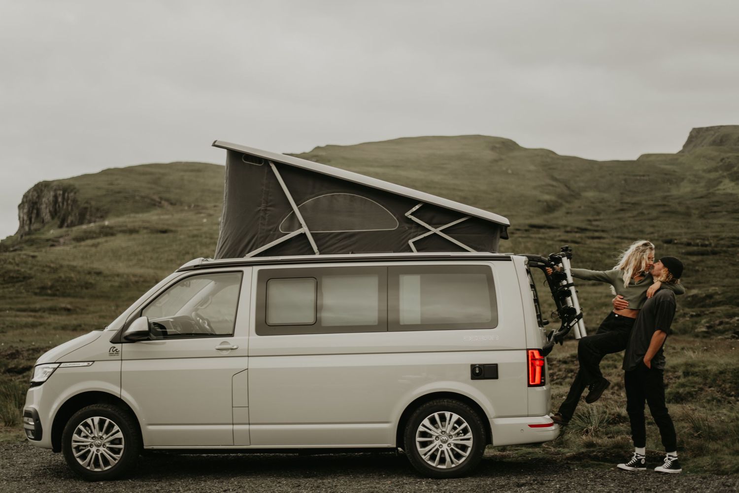 A silver Volkswagen camper van with pop-up roof parked in front of rugged mountain landscape in moody weather.