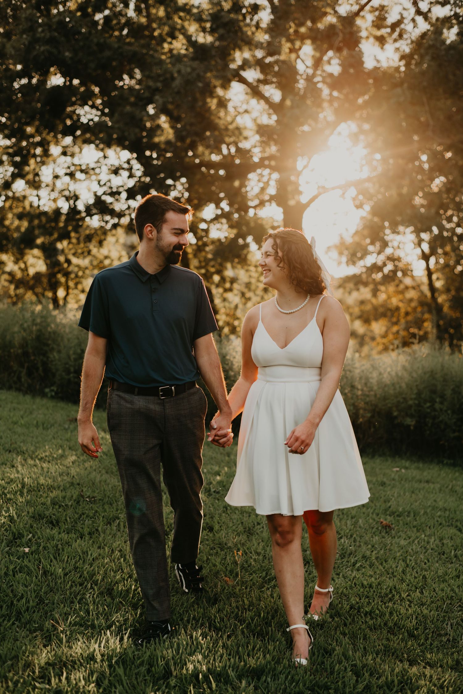 A couple walks hand in hand through a sunlit meadow at golden hour as warm sunlight filters through the trees.