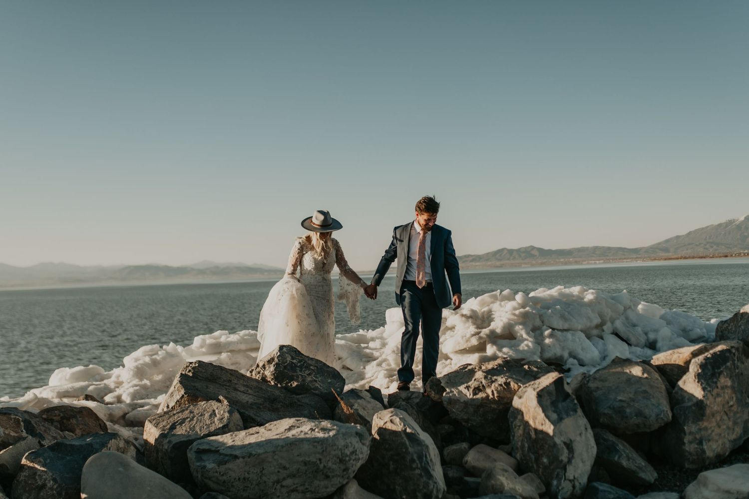 Newlyweds hold hands while standing on rocky jetty by calm waters in Utah.
