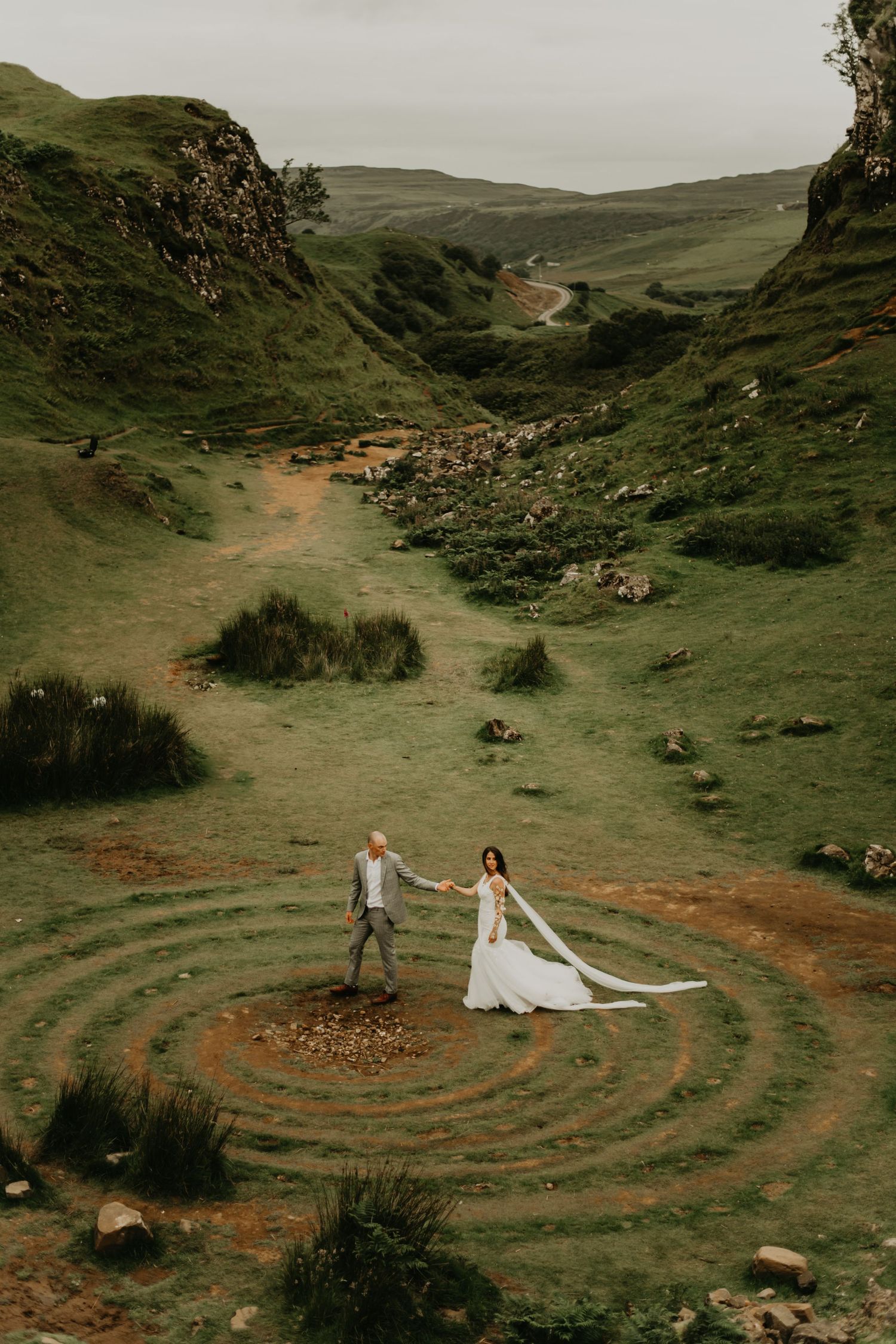 Aerial view of newlyweds standing in circular formation on winding dirt path through green valley in Isle of Skye Scotland.