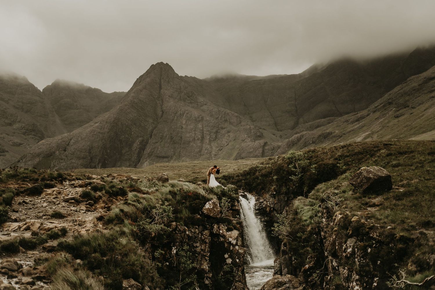 A couple poses near a majestic waterfall in a rugged mountain landscape in isle of Skye Scotland.