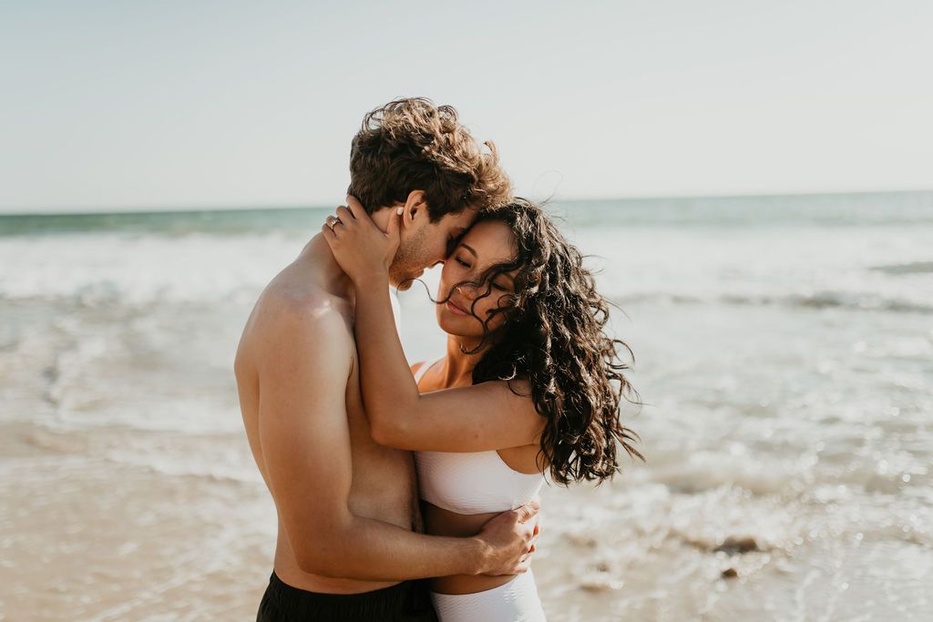 A loving couple embraces romantically on a sun-drenched beach with ocean waves in the background.