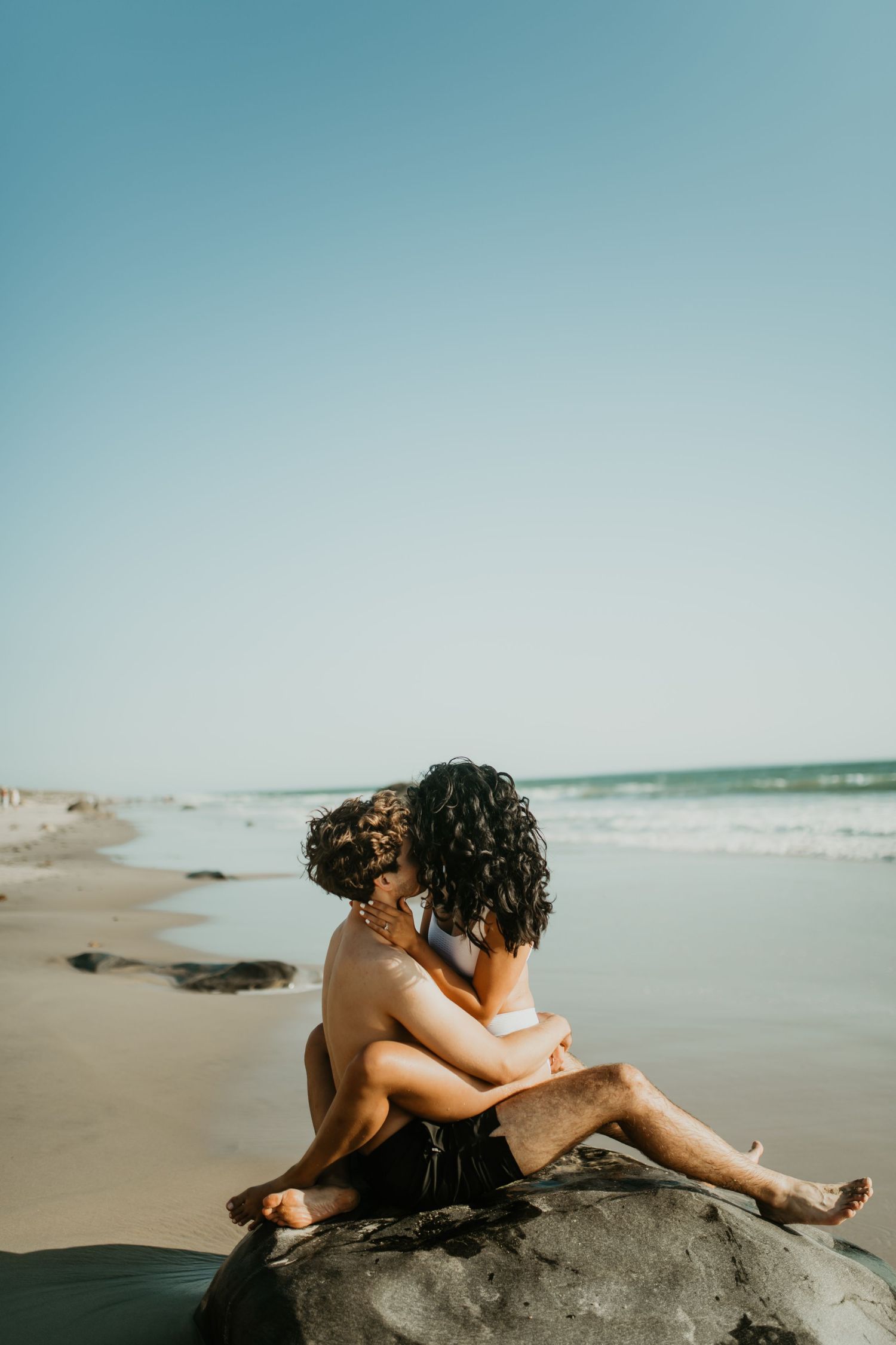 Couple embracing on a rock at the beach while waves crash against the shoreline at sunset.